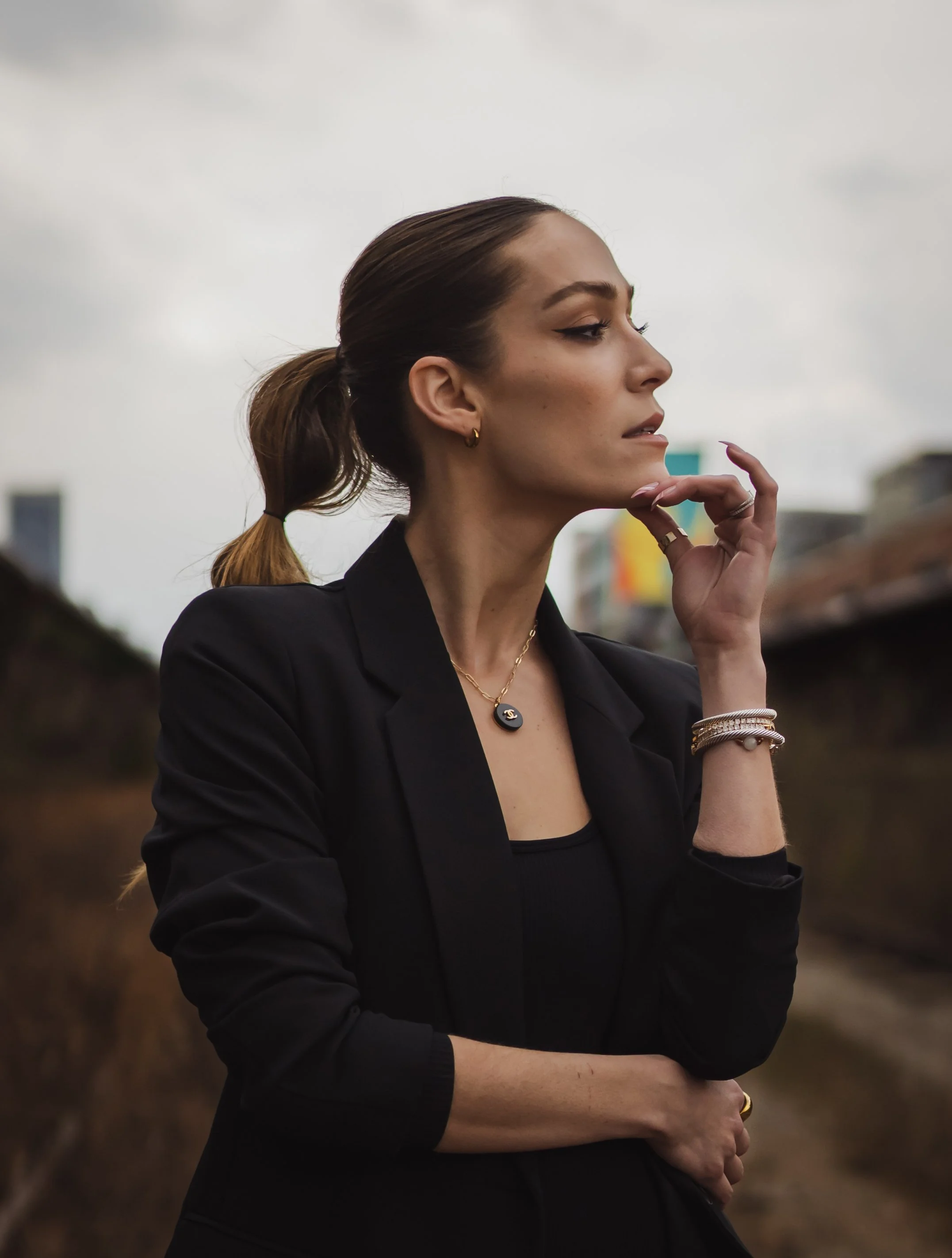 A woman with brown hair in a ponytail, wearing a black blazer and jewelry, stands outdoors under cloudy skies with a contemplative expression.