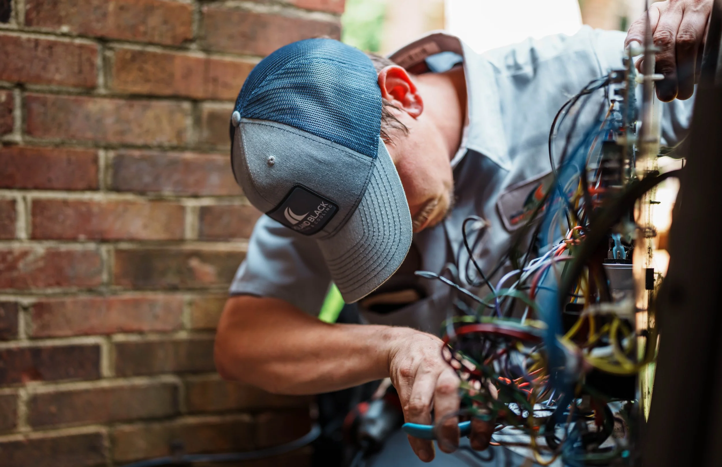 A man wearing a blue and grey cap working on wiring inside a panel or electrical box against a brick wall.