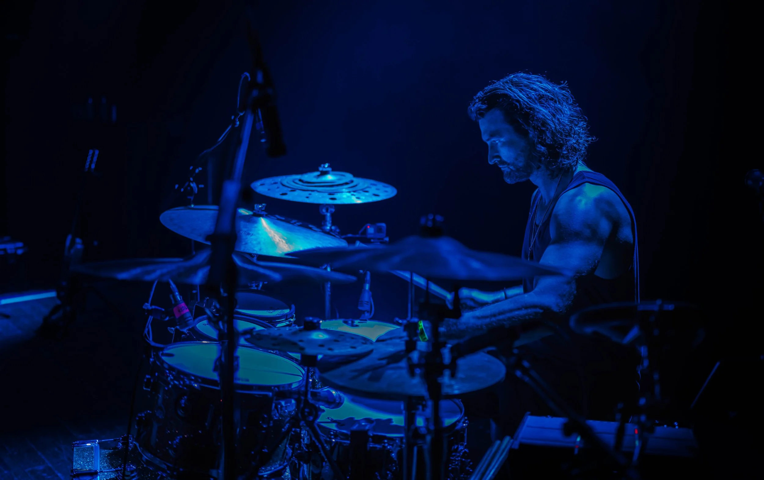 A male drummer with long curly hair playing a drum set on stage under blue lighting.