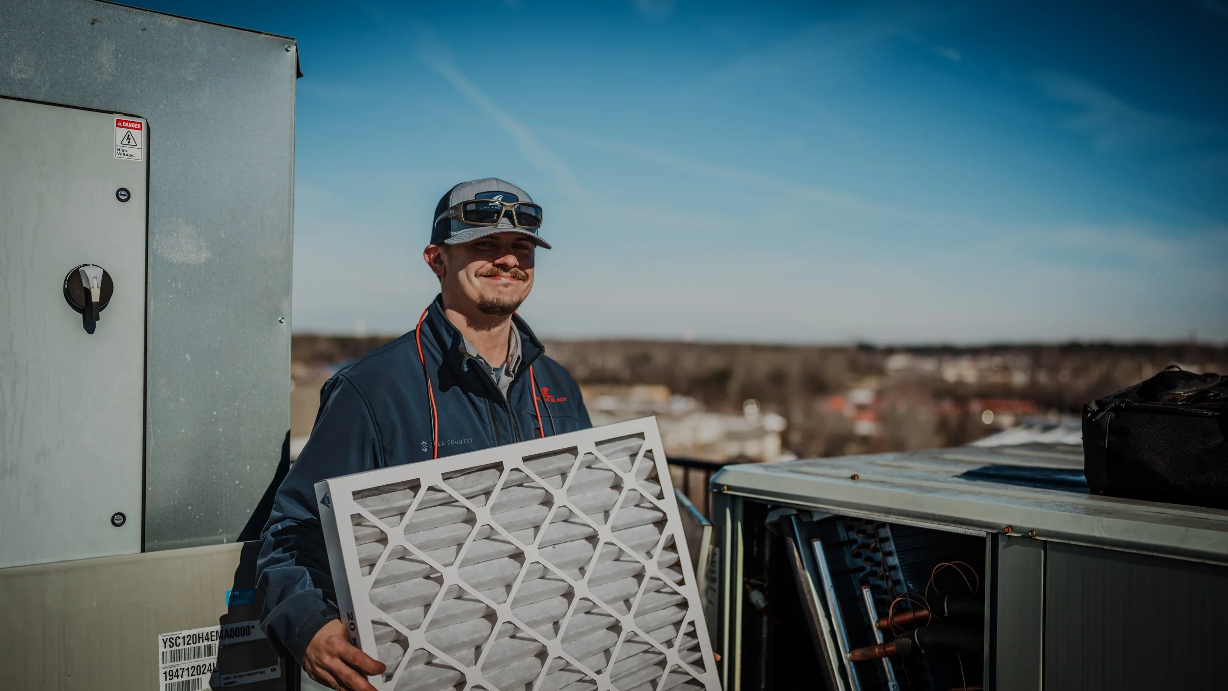 A man wearing a baseball cap and safety glasses holds a large HVAC filter in front of an outdoor air conditioning unit on a rooftop, with a clear blue sky and a cityscape in the background.
