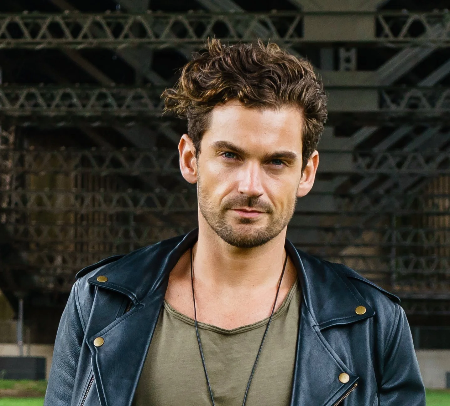 A young man with wavy brown hair, light beard, and blue eyes, wearing a green t-shirt and black leather jacket, standing outdoors in front of a bridge structure.
