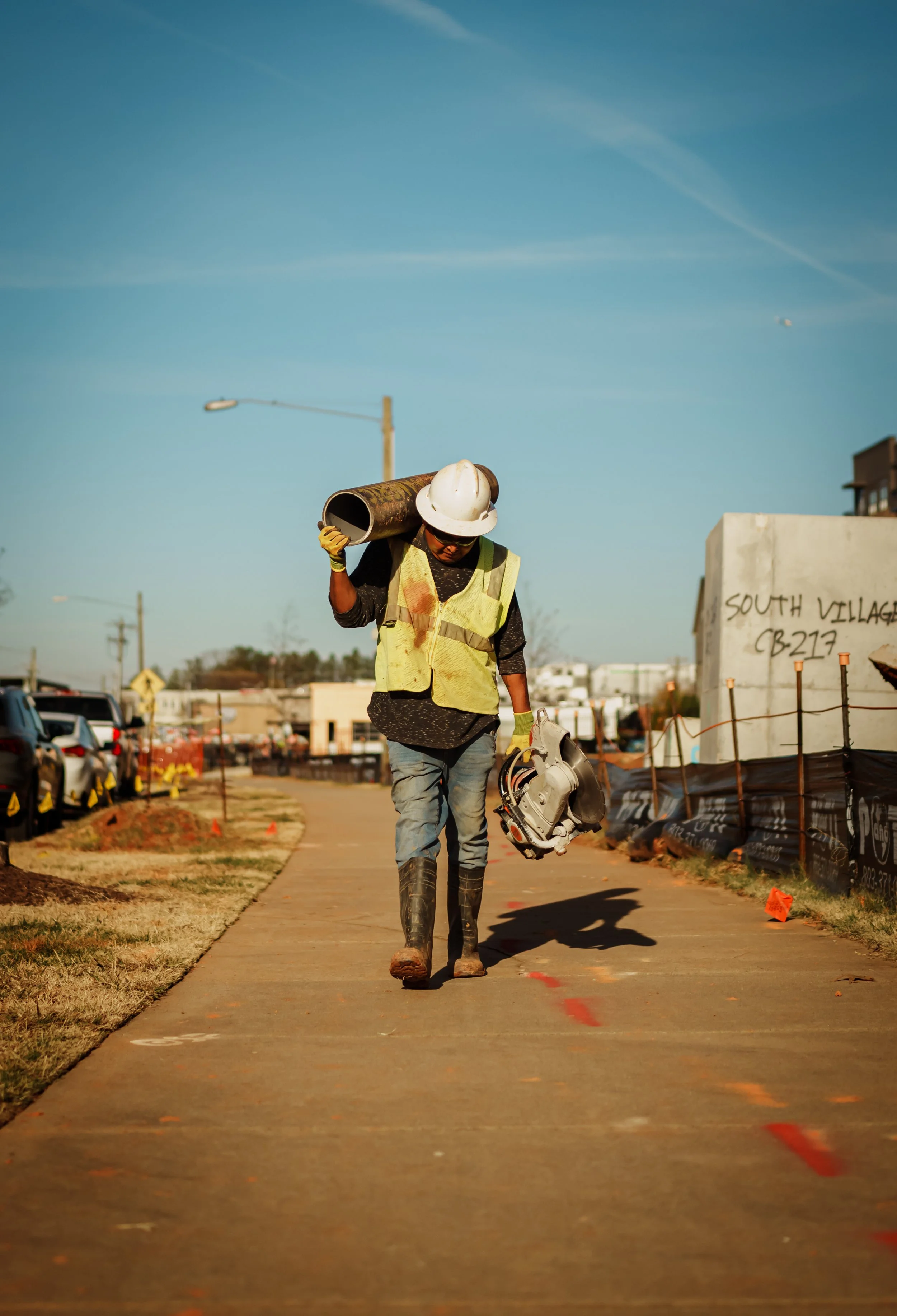 Construction worker wearing a white helmet, sunglasses, and yellow safety vest carrying a duct pipe and a power saw while walking on a sidewalk at a construction site.