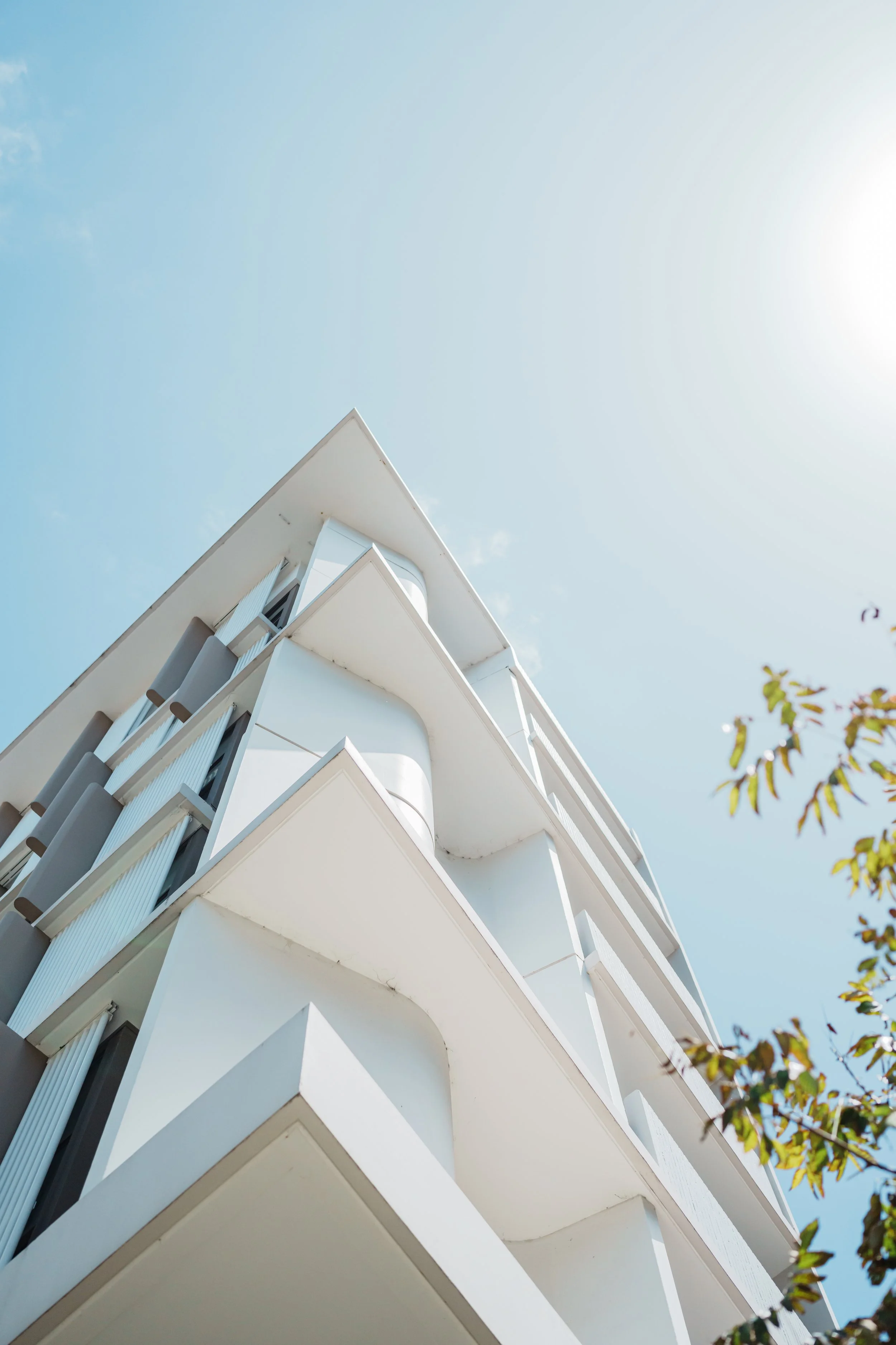 A modern white multi-story building with balconies, seen from below against a clear blue sky.
