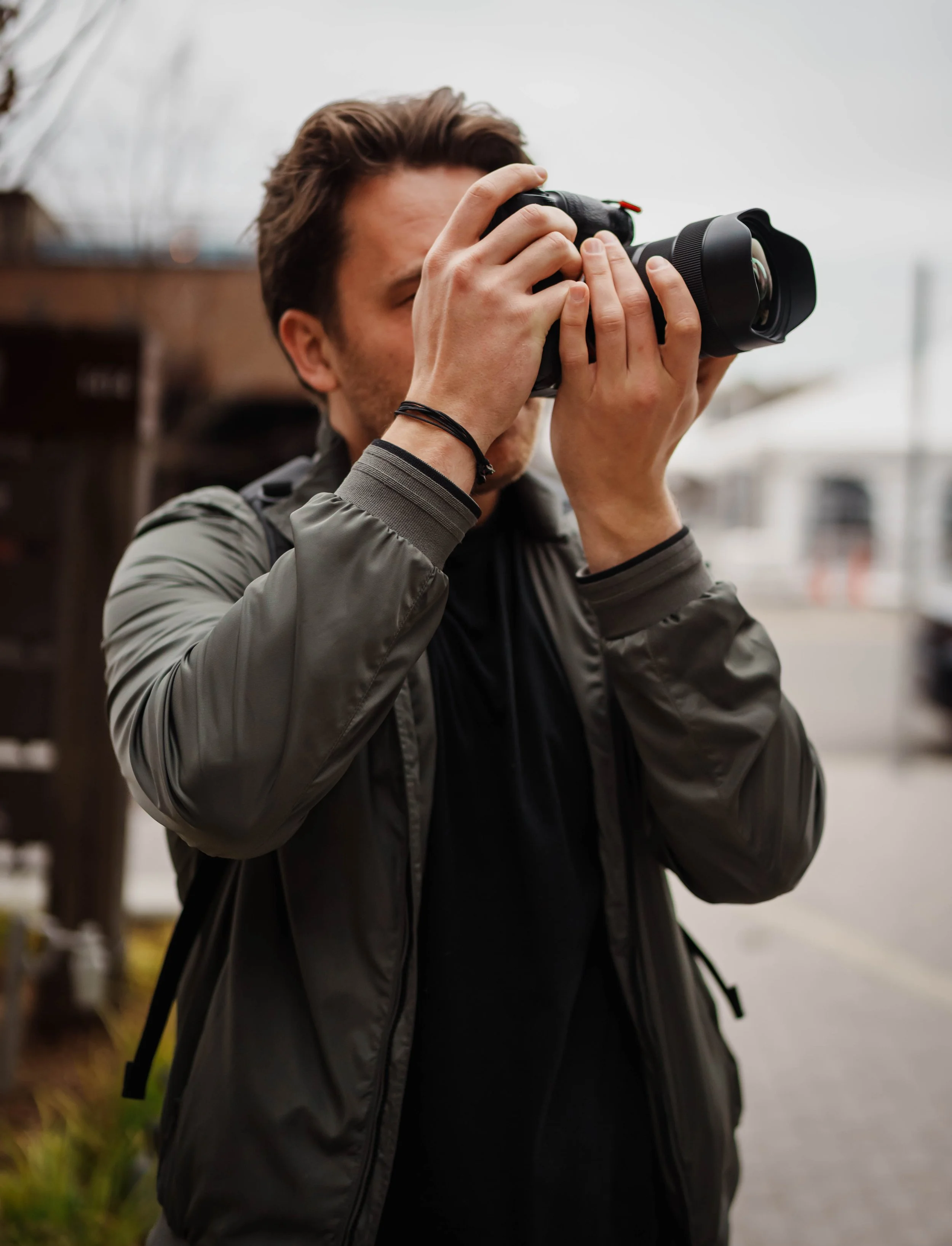 A man taking a photograph in an outdoor urban setting, wearing a gray jacket and holding a professional camera to his eye.