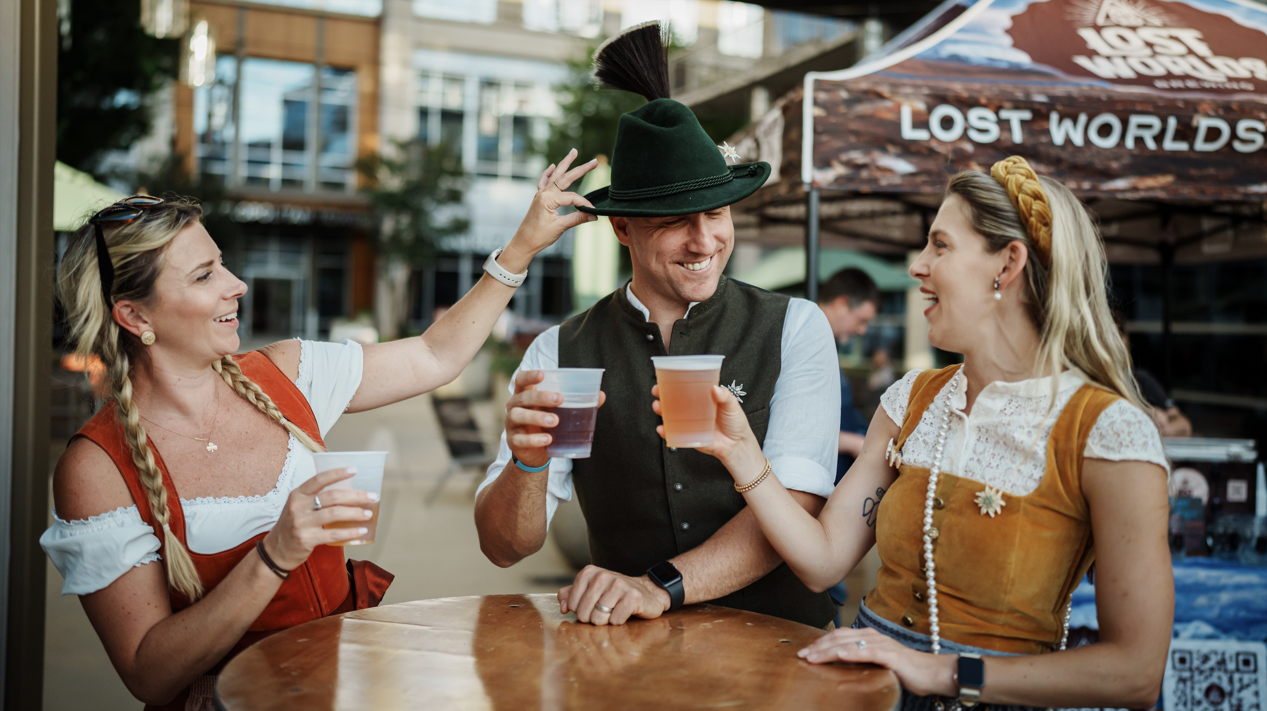 Three people dressed in traditional German attire celebrating outdoors at a table, toasting with drinks, with a man wearing a green hat. A banner with the words "Lost Worlds" is in the background.