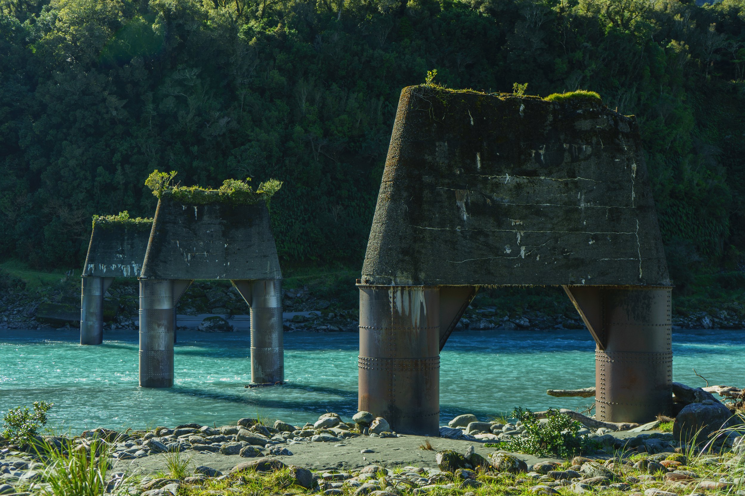 Two large abandoned bridge supports in a river with forested hills in the background.