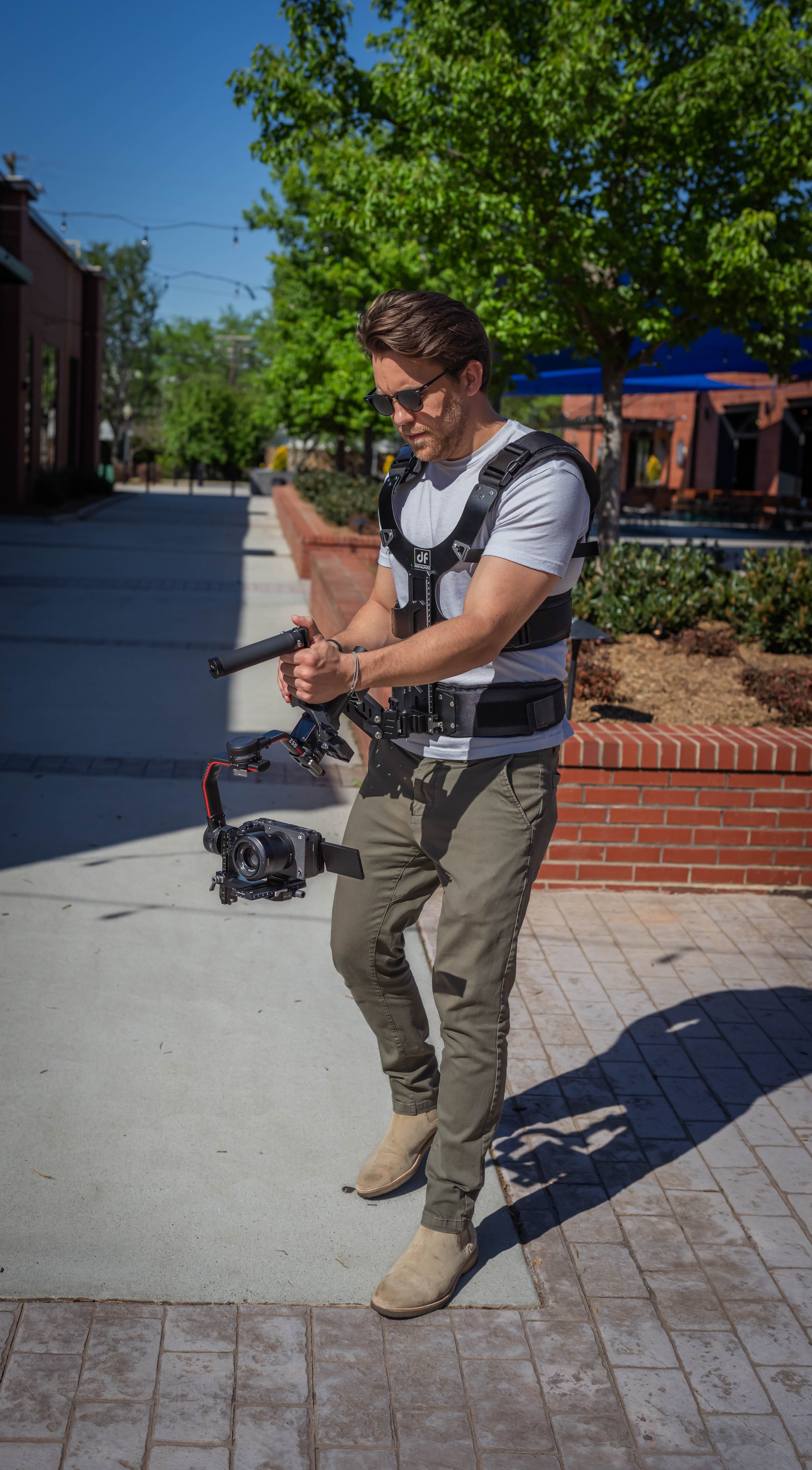 A man wearing sunglasses and casual clothes, holding a camera stabilizer with a mounted camera, standing outdoors on a sunny day with trees and buildings in the background.
