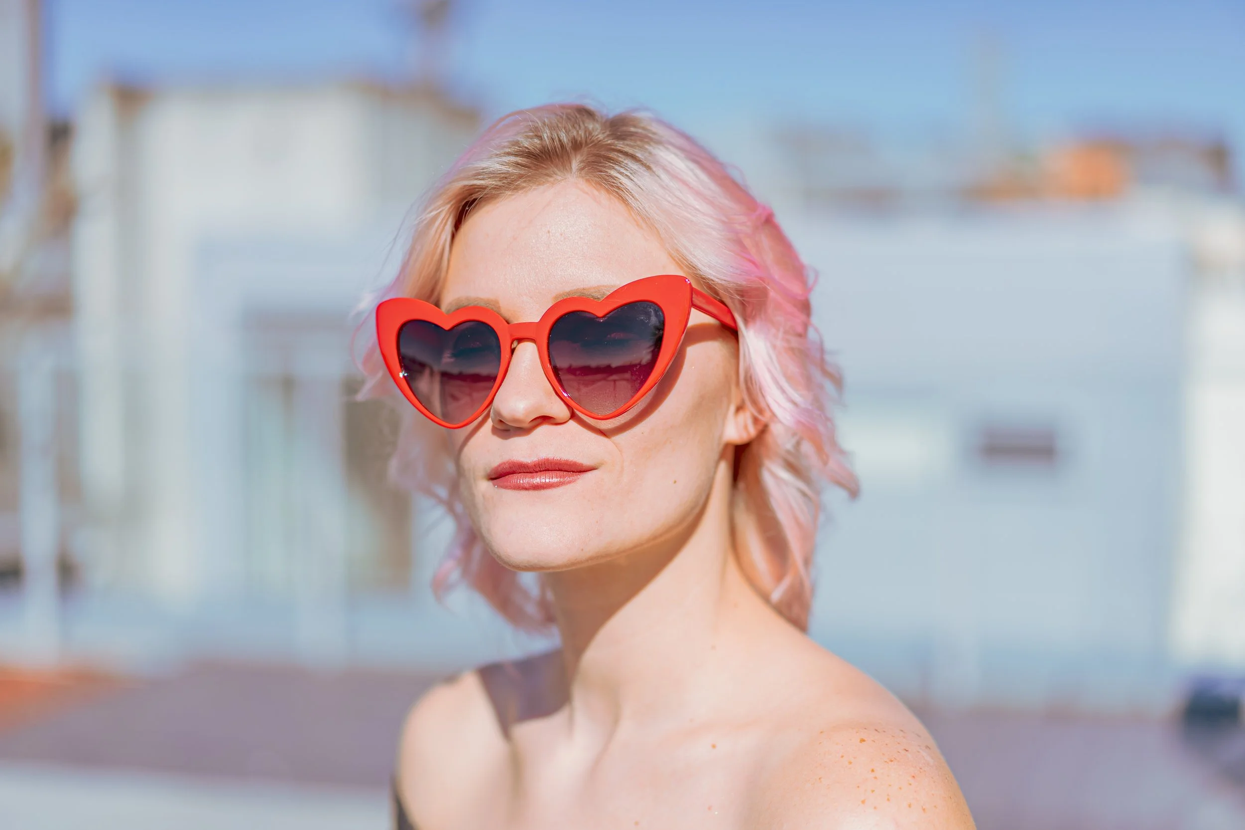 Woman with pink and blonde hair wearing red heart-shaped sunglasses outdoors with a modern building in the background.