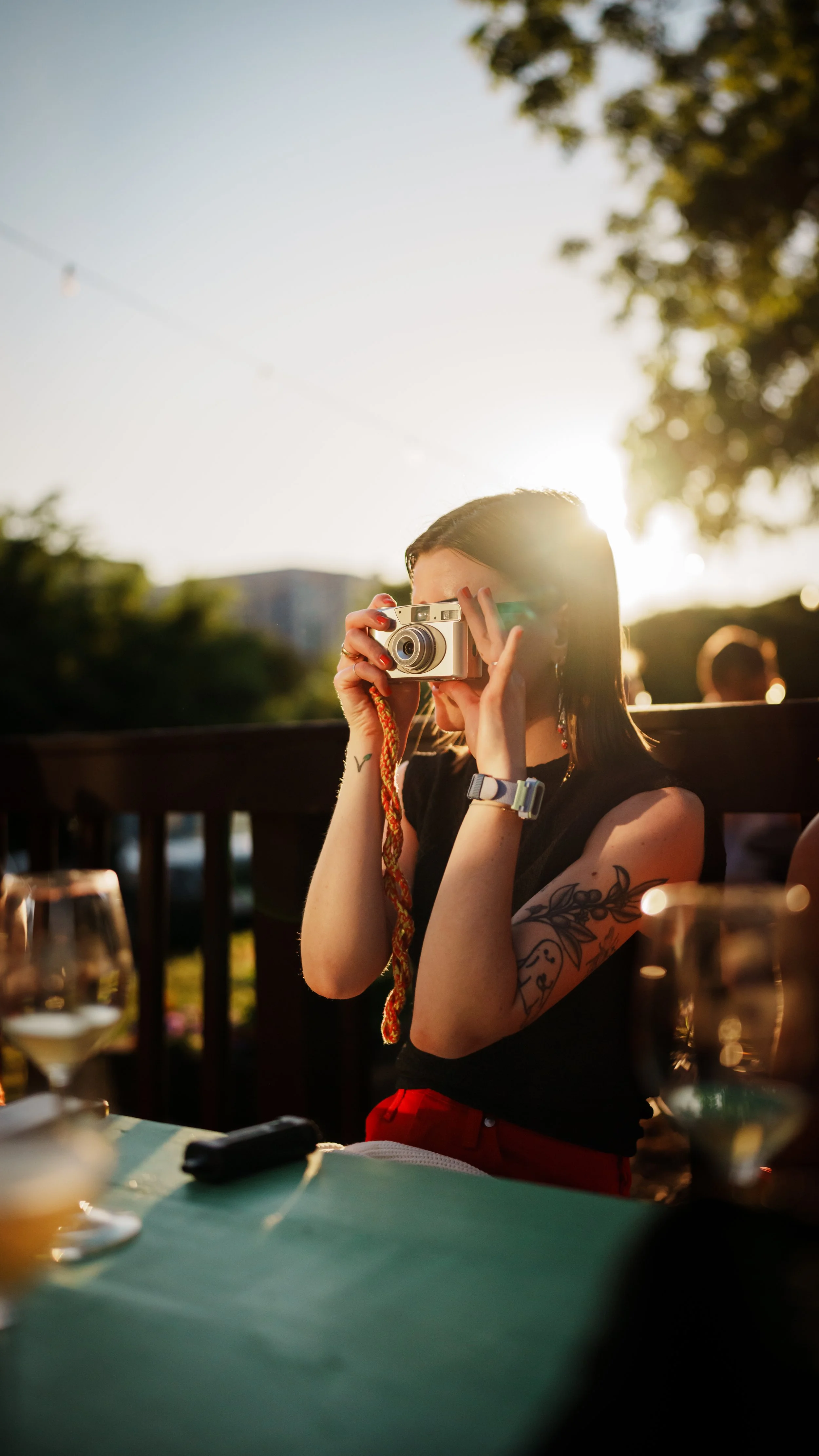 Woman with tattoos taking a photo with a vintage camera at an outdoor gathering during sunset.