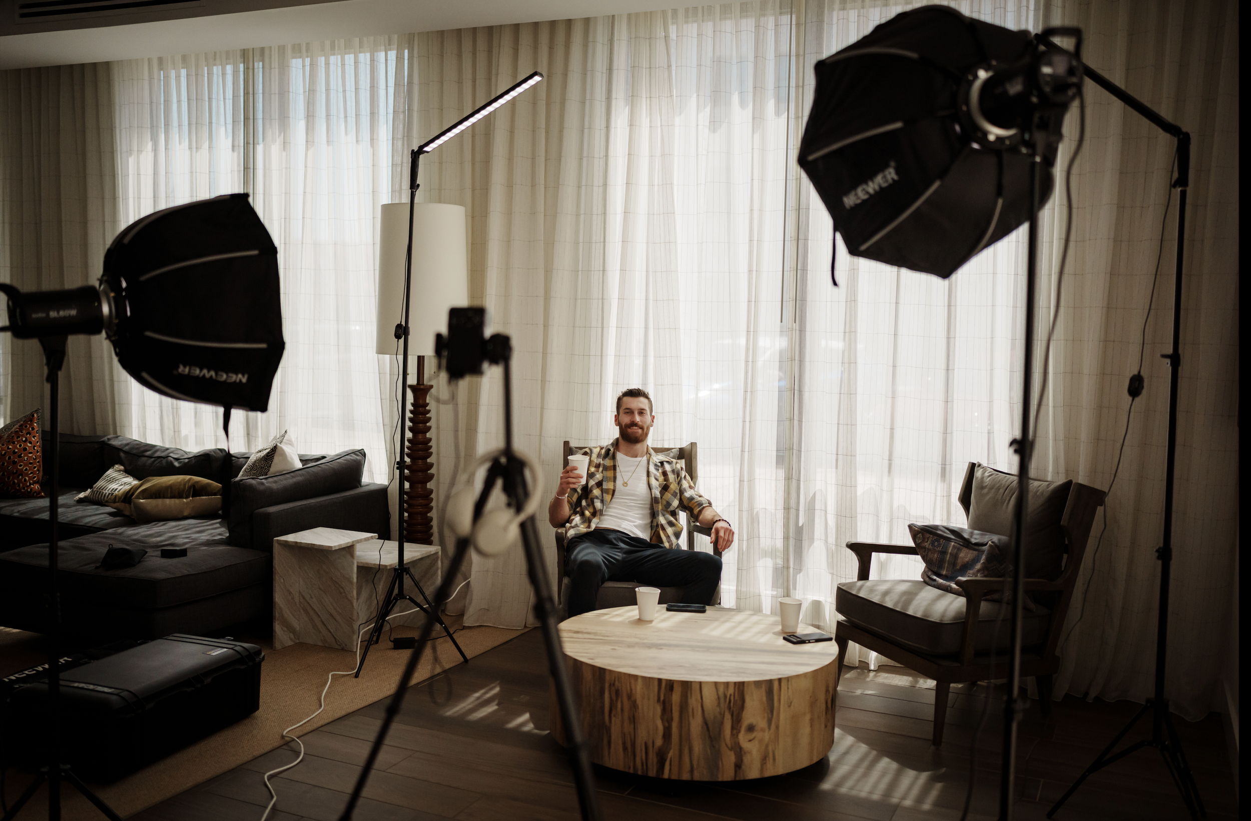 Man sitting in a chair, smiling, holding a drink, surrounded by film lighting equipment in a brightly lit living room with curtains.