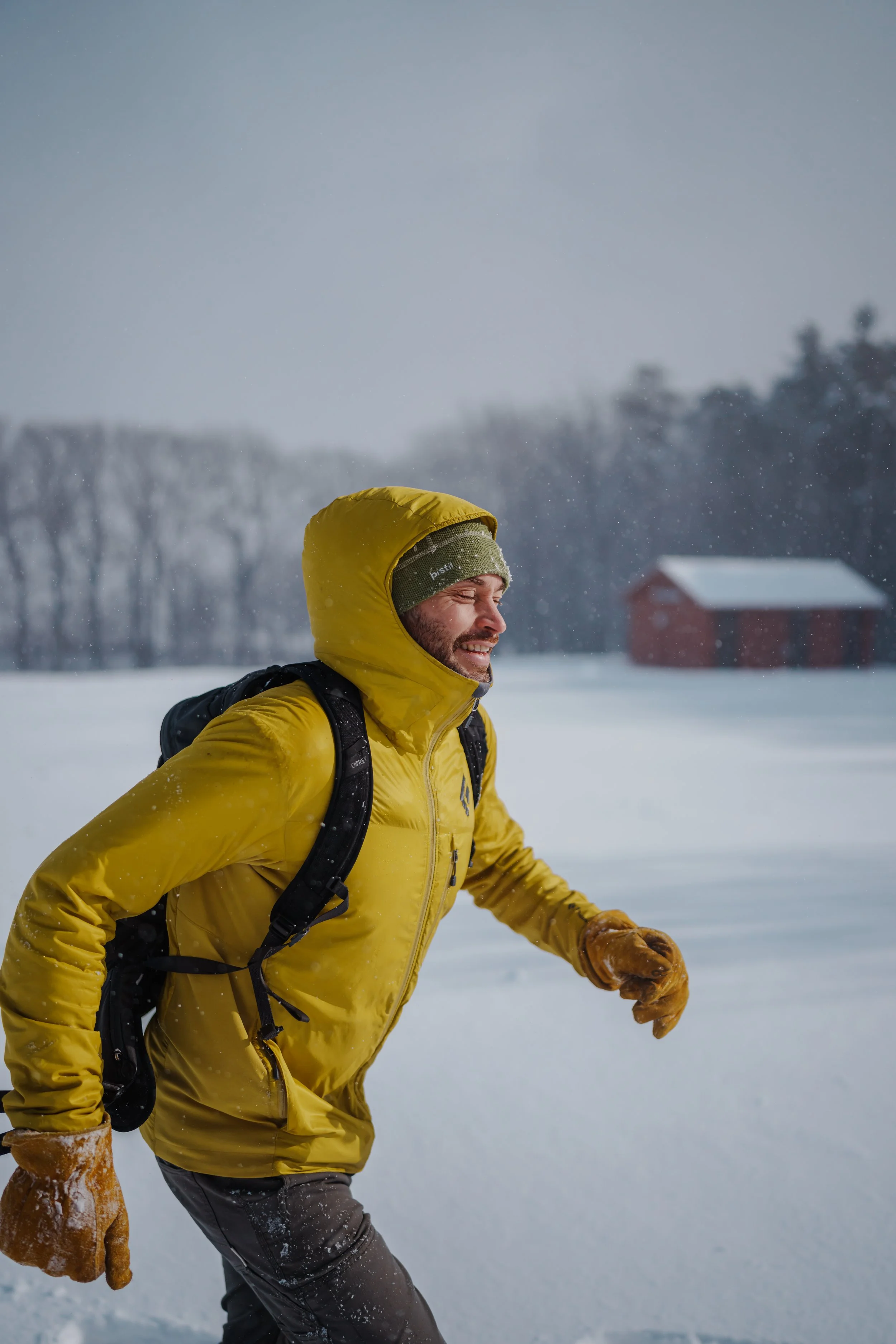A man in a yellow winter jacket running through snow with a backpack and brown gloves, smiling with snow falling.