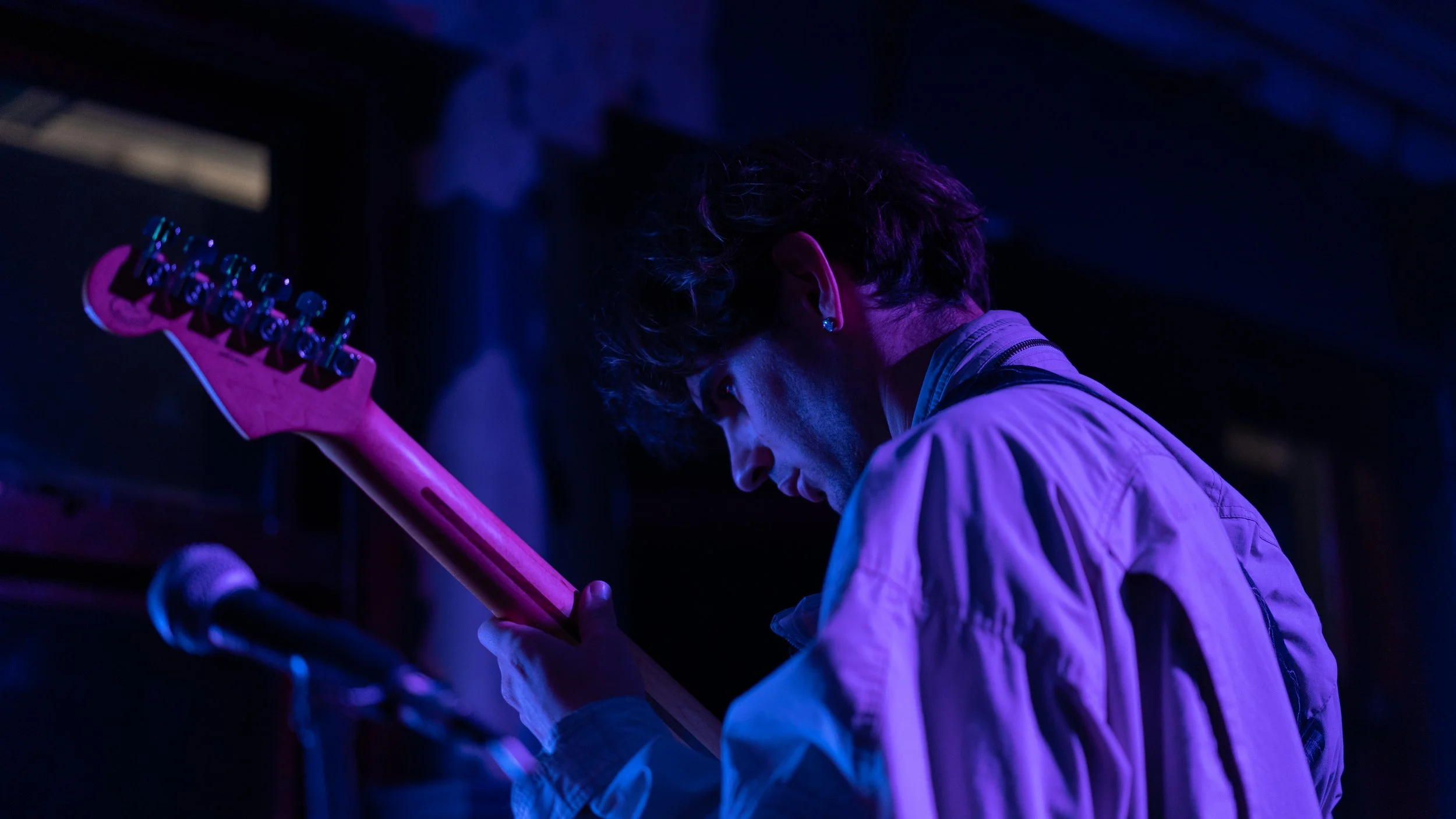 A musician in a light-colored jacket playing an electric guitar under purple and blue stage lights, with a microphone in the foreground.