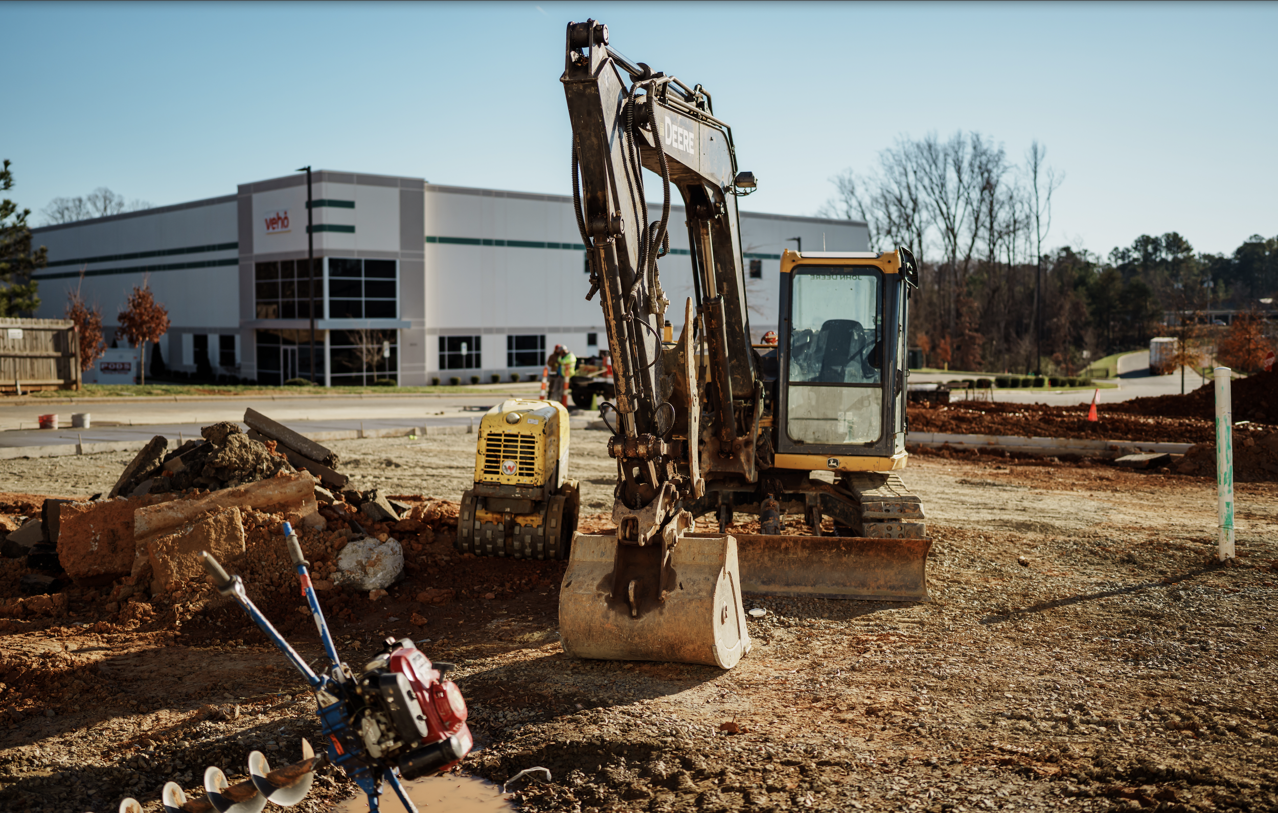 Construction site with a small excavator operating on dirt, construction tools on the ground, and a modern building in the background under a clear blue sky.