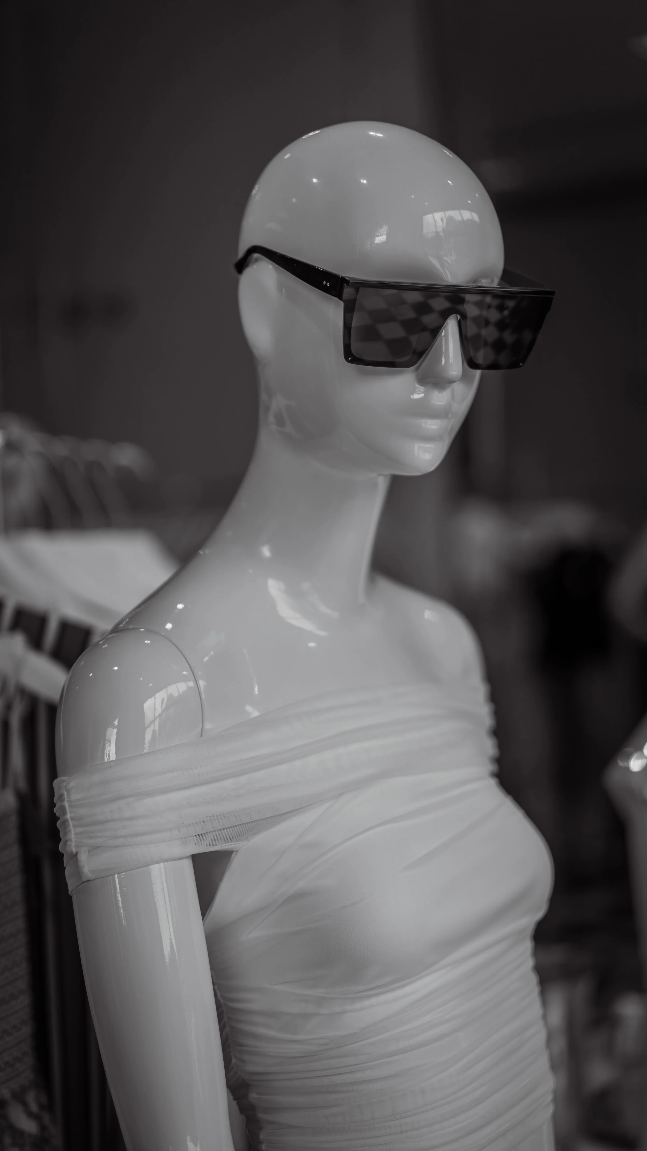 A white mannequin wearing large black sunglasses and a off-the-shoulder, ruched white dress.