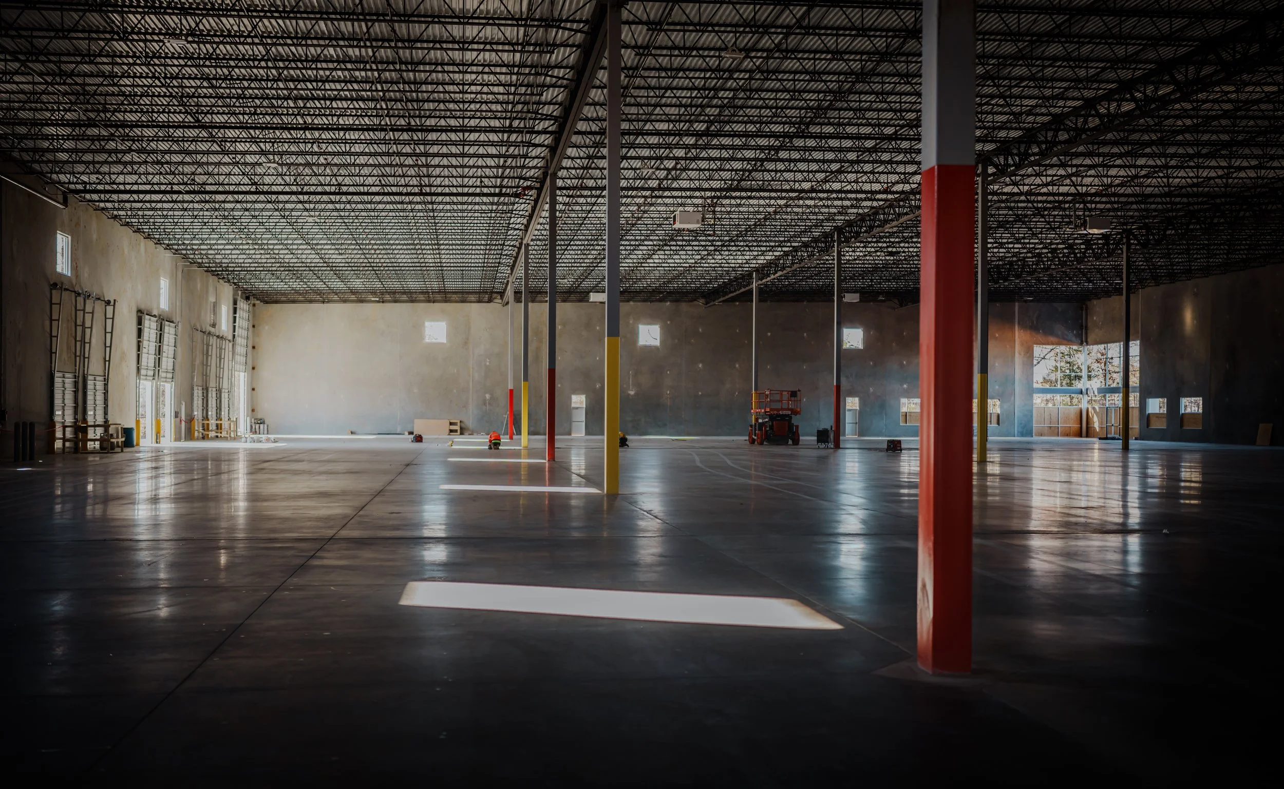 Empty industrial warehouse with high ceiling and support columns, sunlight streaming through windows, construction equipment scattered on the polished concrete floor.