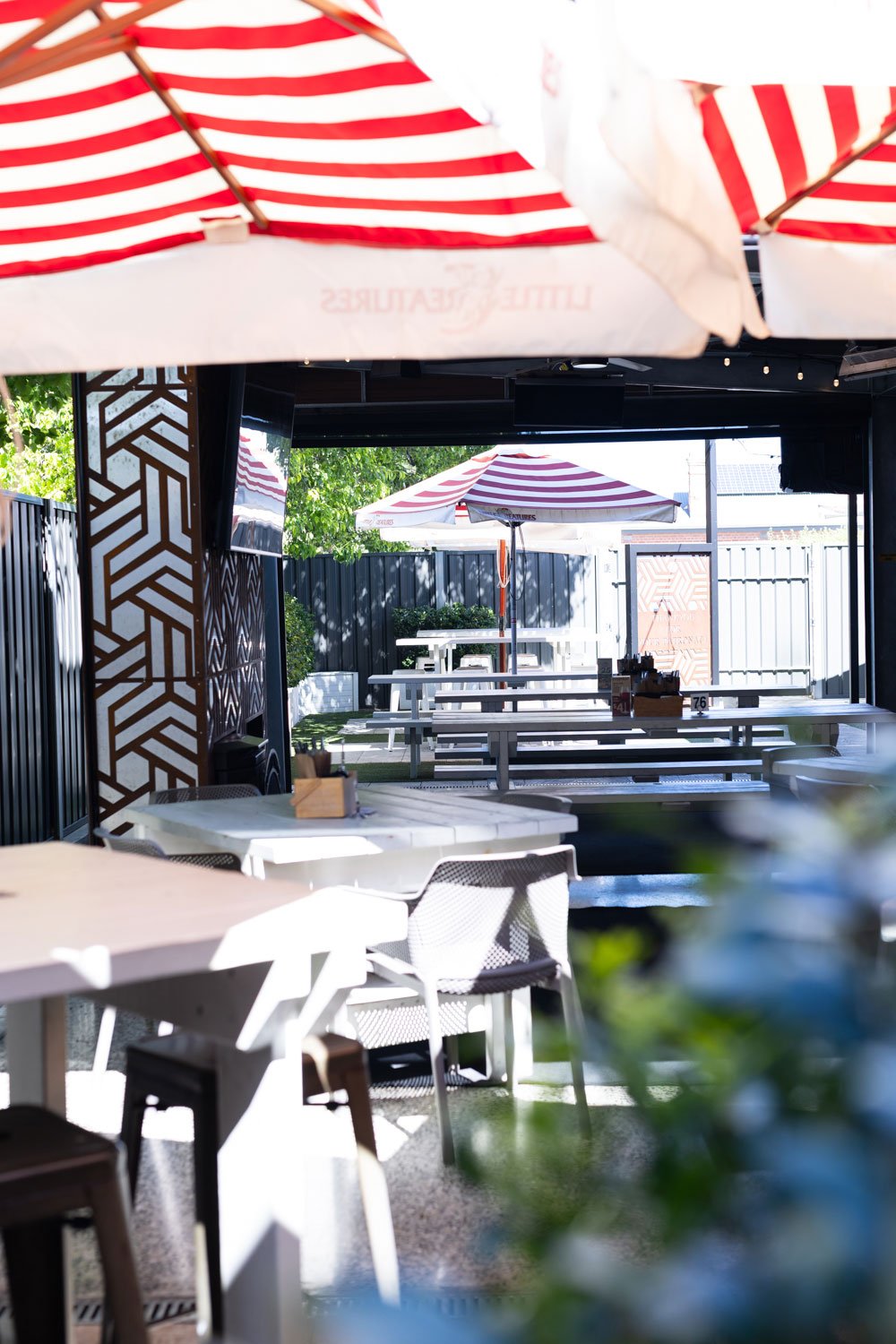 Outdoor patio with white tables, stools, and picnic benches under red and white striped umbrellas, with string lights overhead, and a small bar area in the background.