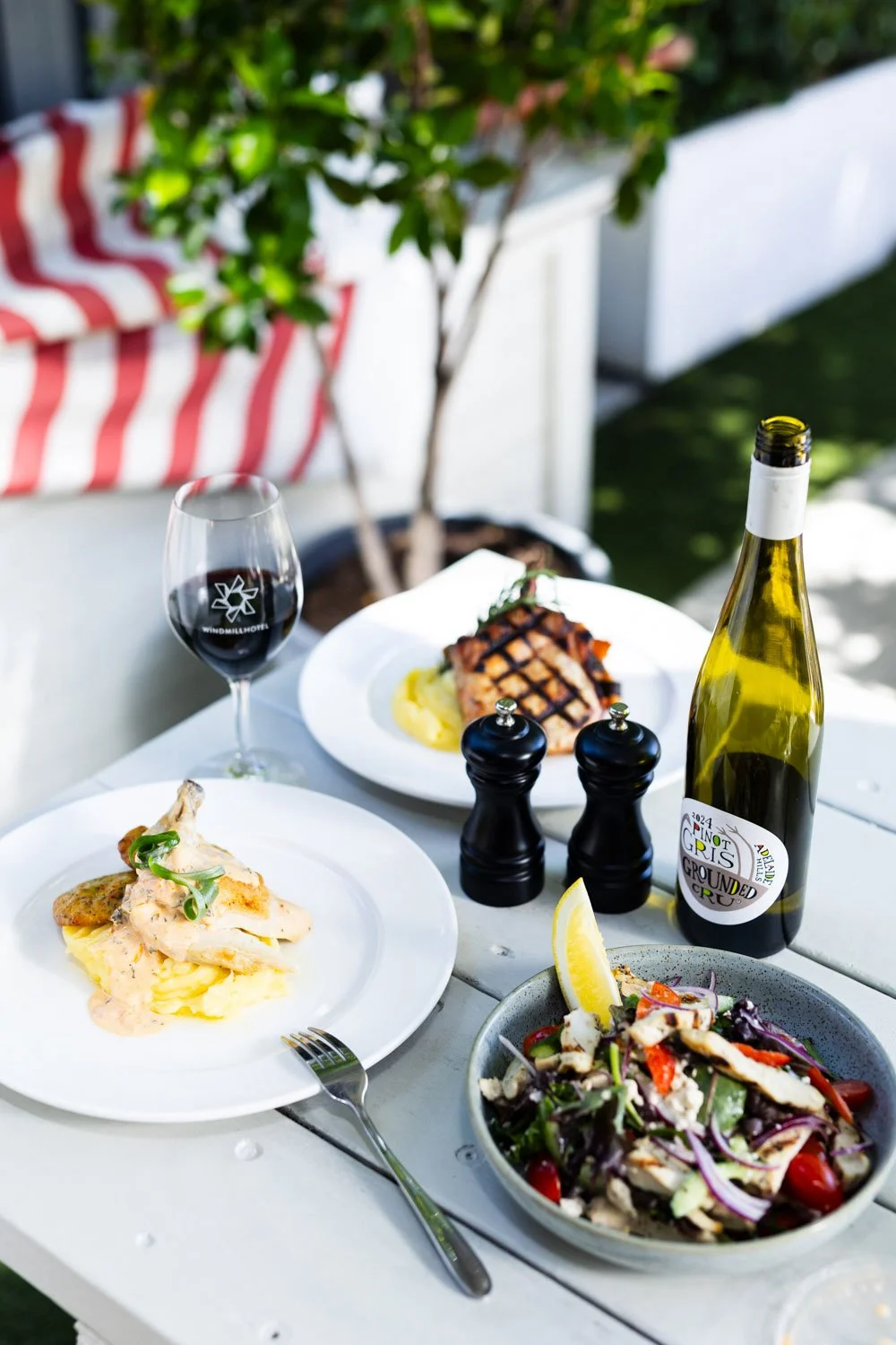 A cheese platter with various cheeses, fresh strawberries, and garnishes on a black plate, accompanied by a glass of white wine and a glass of red wine on a wooden table. There is a basket of assorted sliced bread, a small gold-colored container, and a vase with pink flowers in the background.