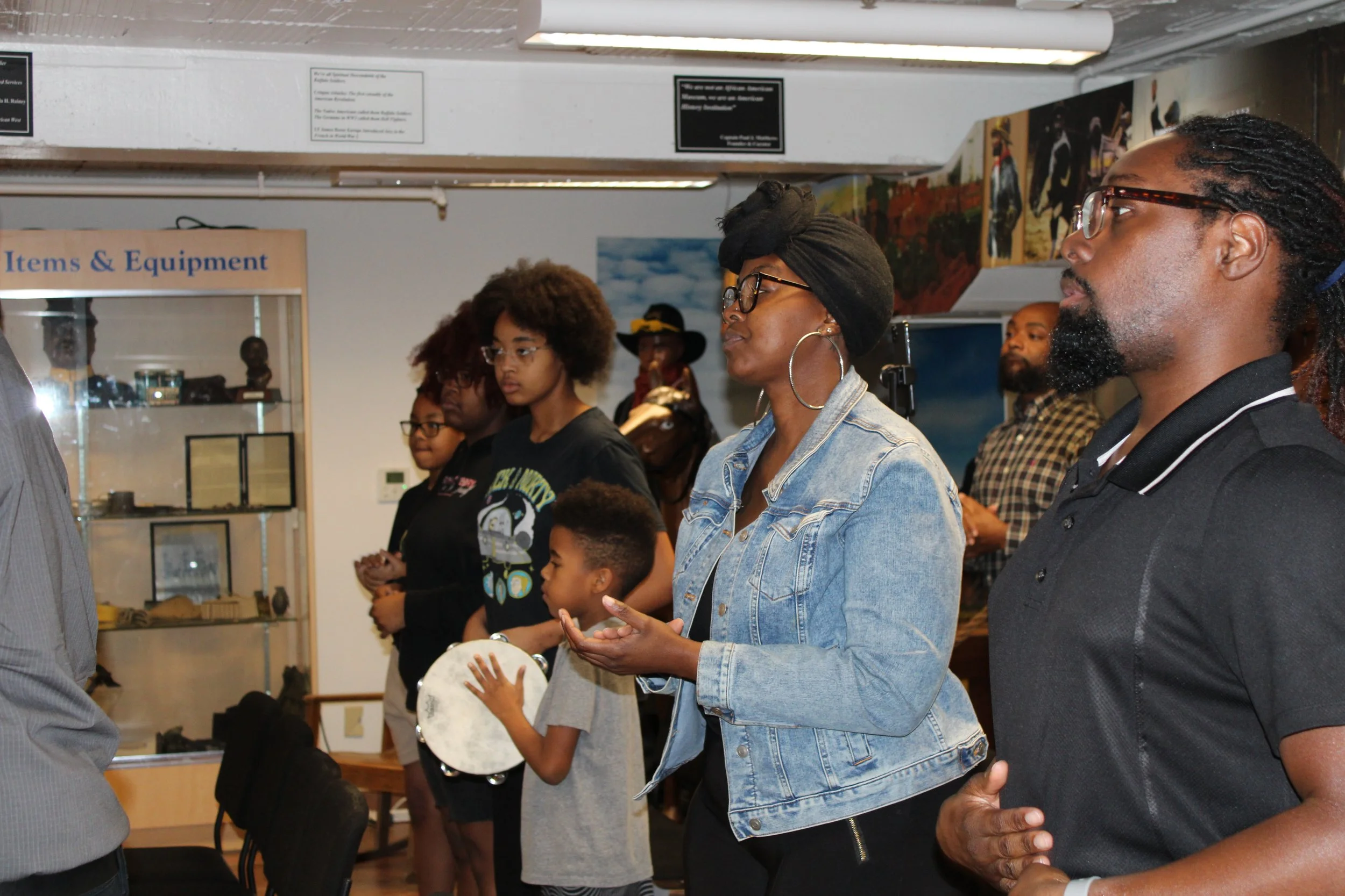 Group of people standing in church, some holding musical instruments, in a room with display cases and artwork on the walls.