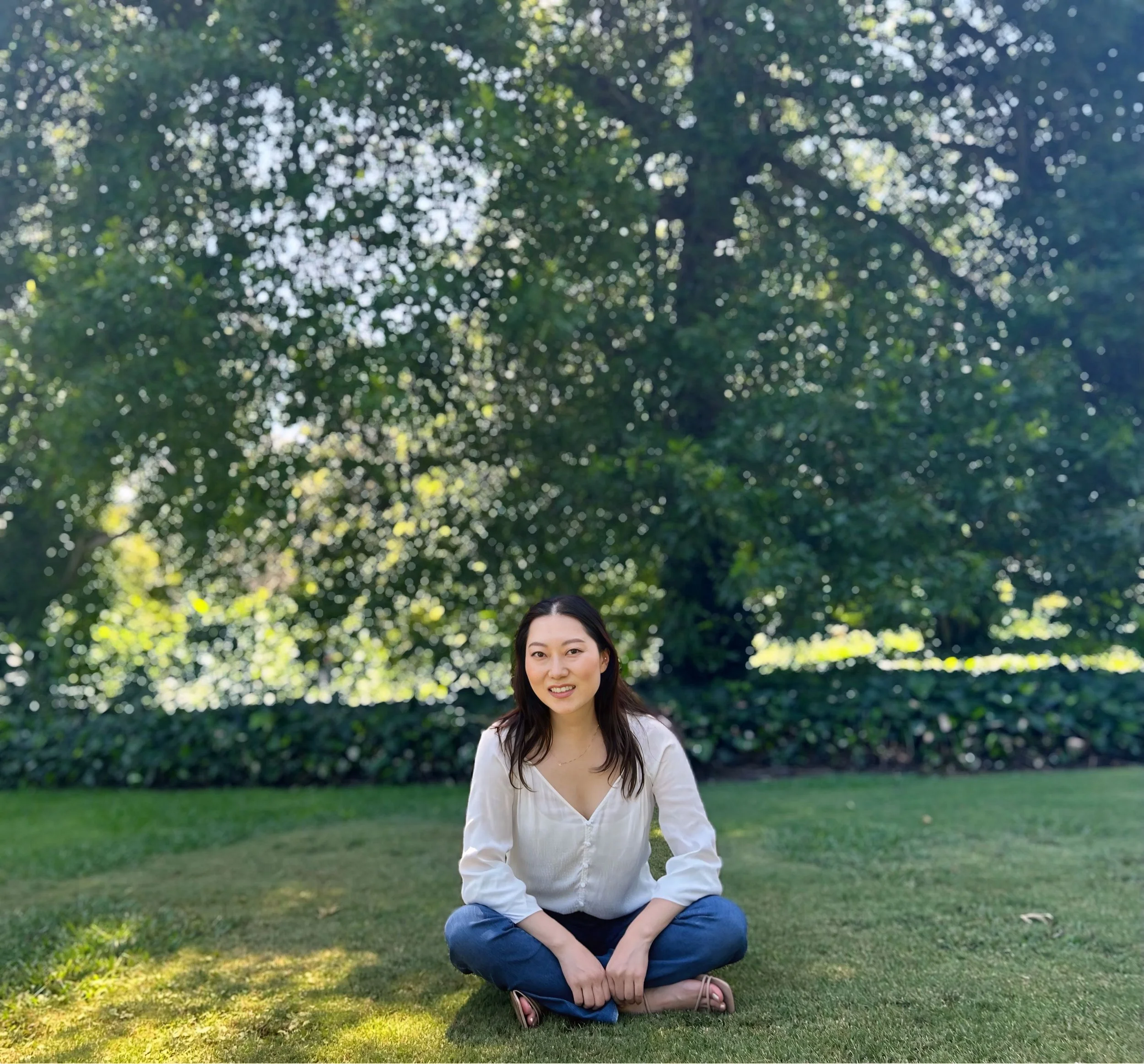 Woman sitting cross-legged on grass in a park, smiling, with lush green trees in the background.