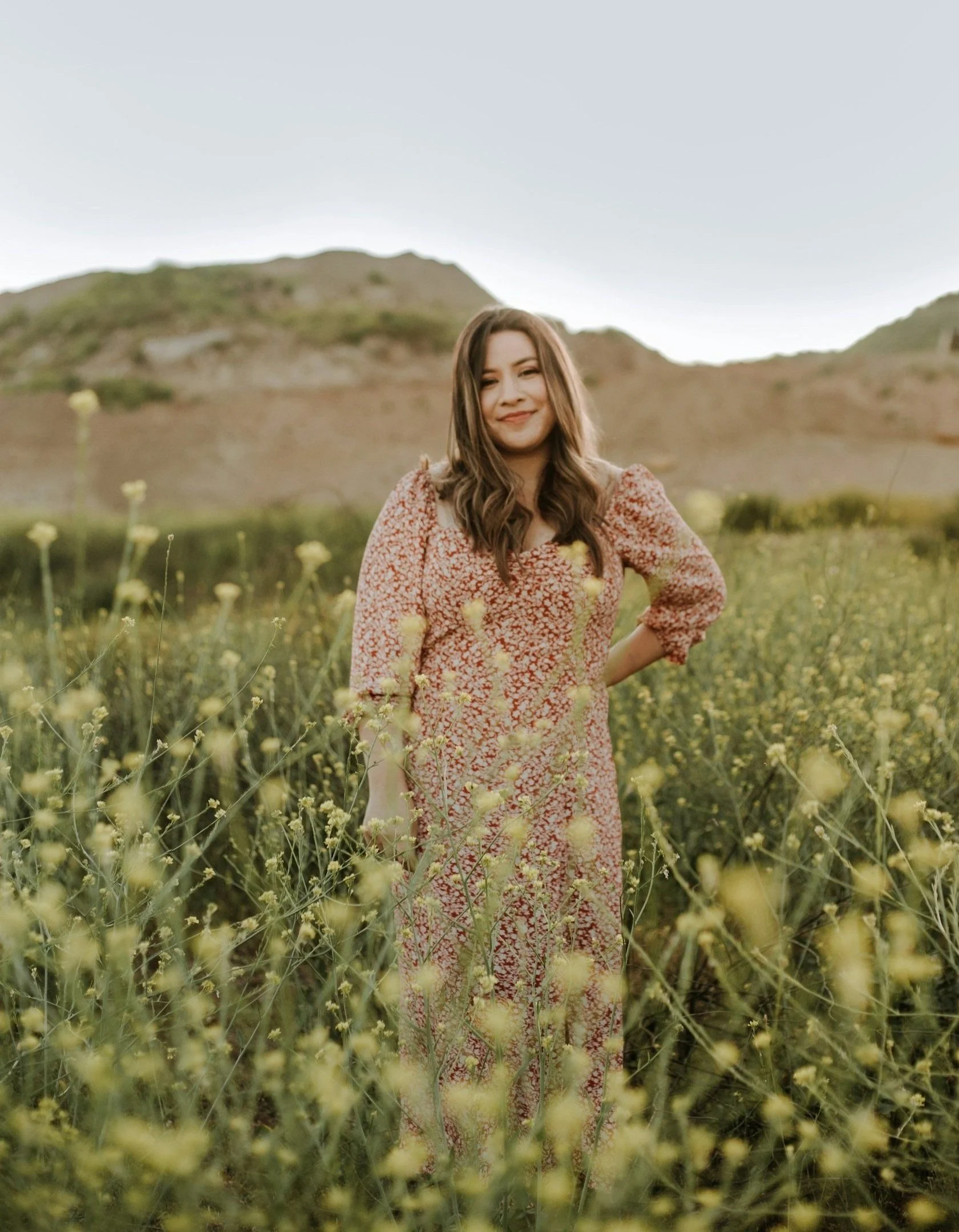 Woman in a long floral dress standing in a yellow flower field with hills in the background.
