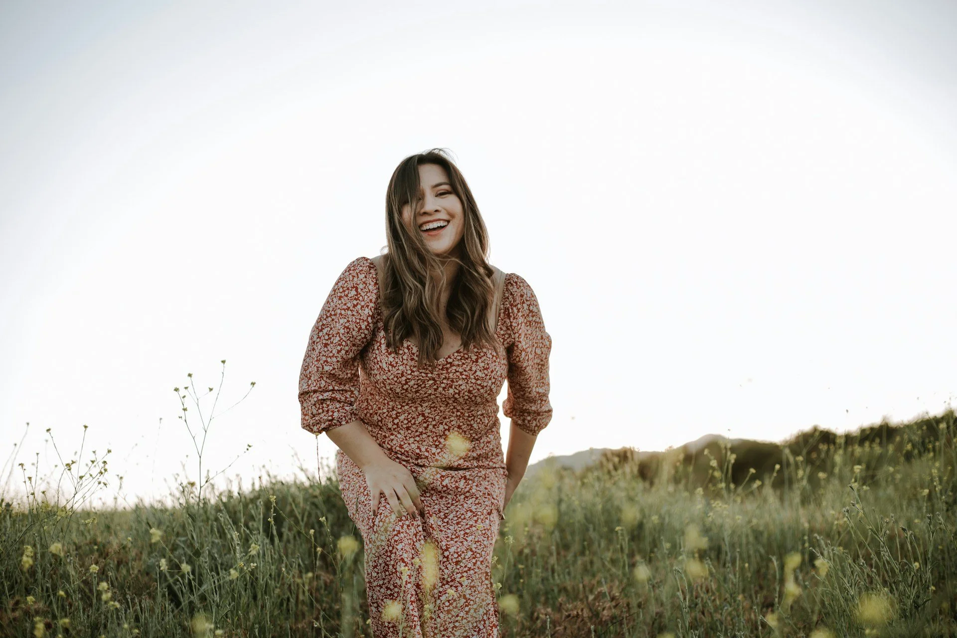 A woman in a patterned red and white dress smiling and laughing outdoors in a grassy field with wildflowers, during daylight.