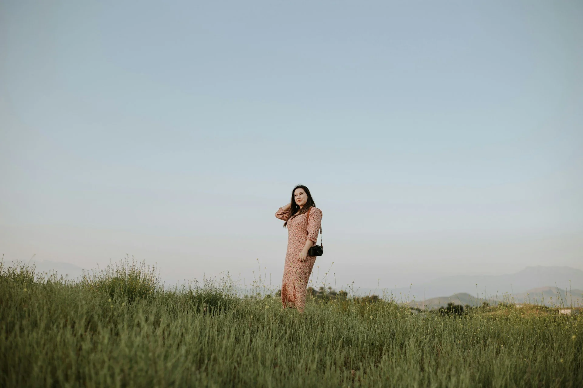 A woman in a floral dress standing in a grassy field with mountains in the distance, holding a camera slung over her shoulder under a clear sky.