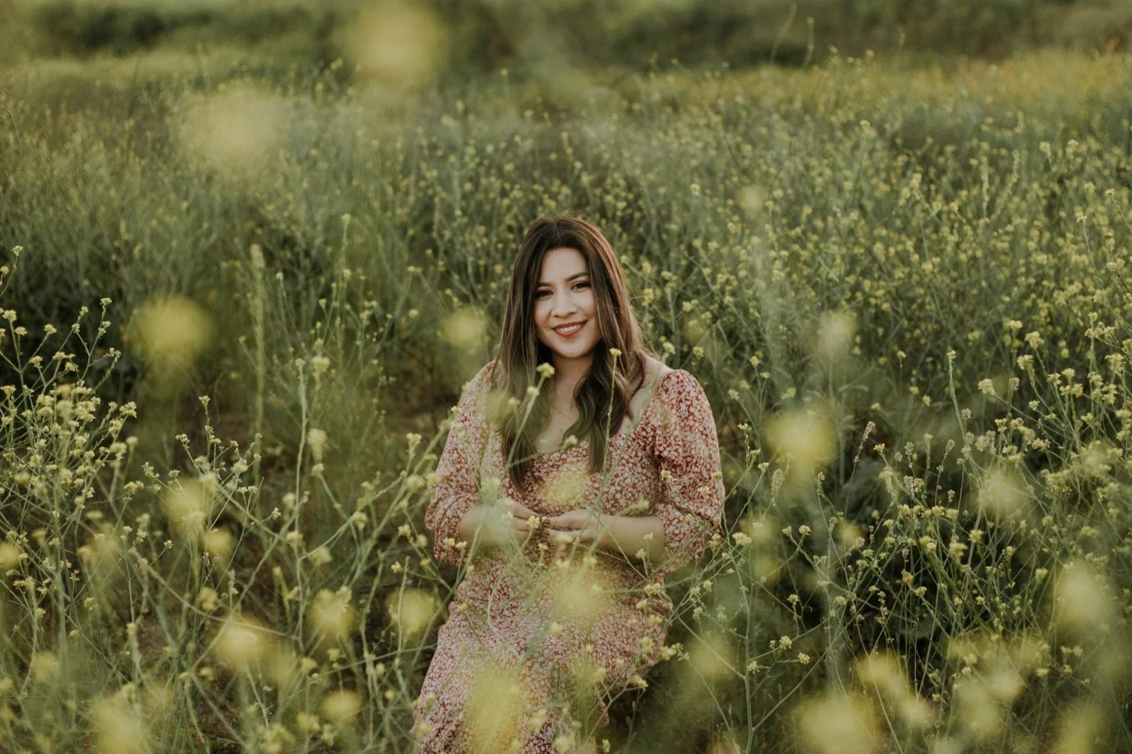 A woman sitting among yellow flowers in a green field, smiling at the camera.