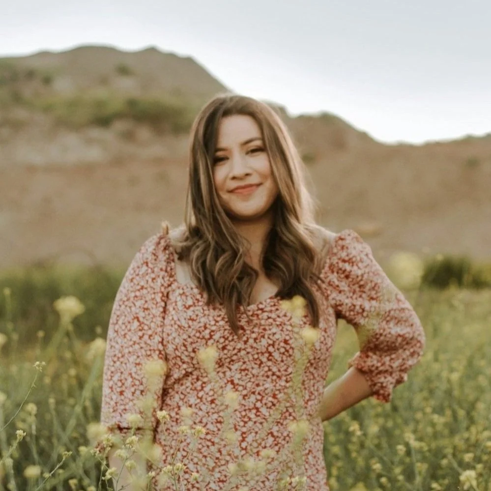 A woman with shoulder-length brown hair wearing a floral red and beige dress standing in a field of yellow flowers with hills in the background.