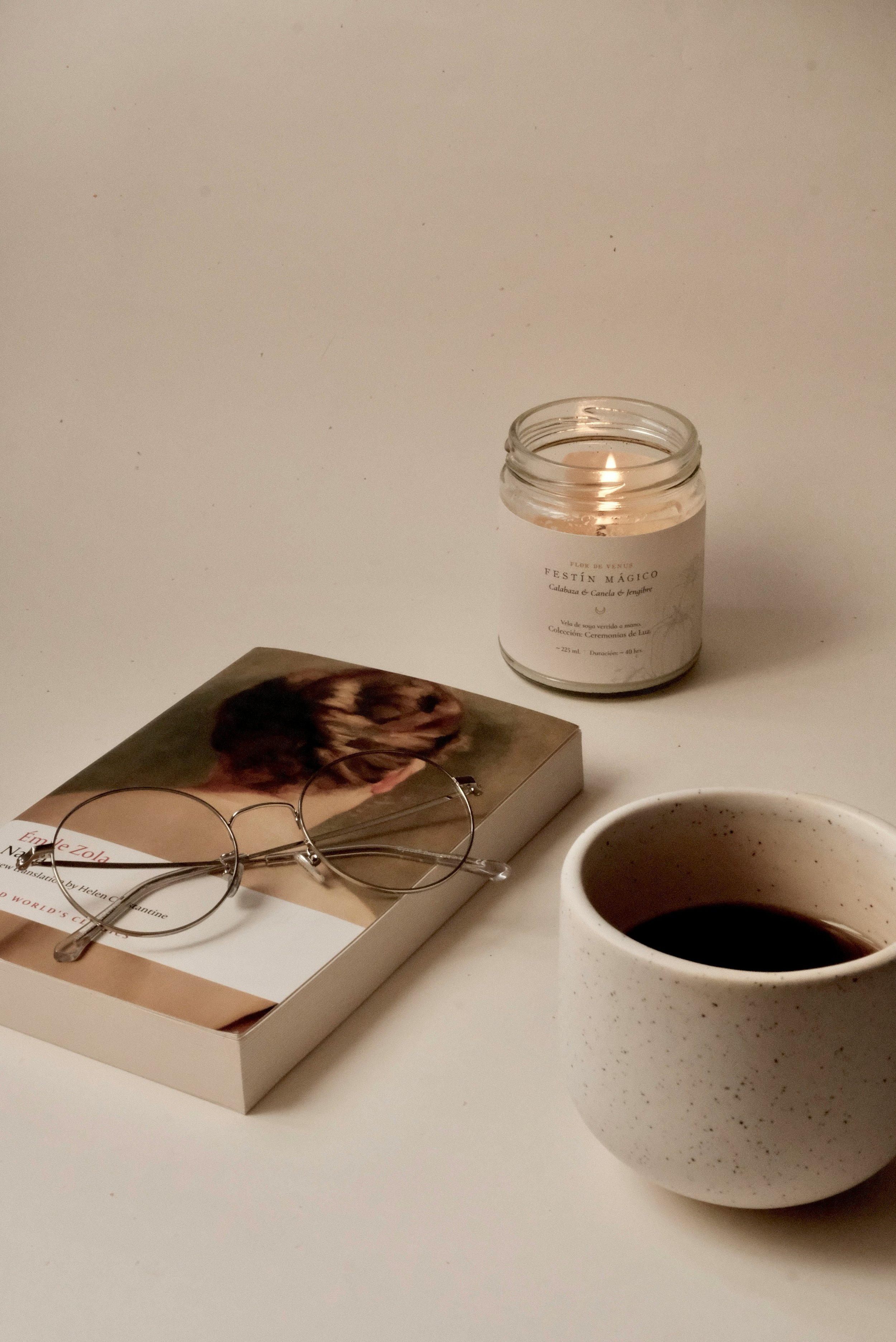 A white table with a book, a pair of glasses resting on the book, a lit candle in a glass jar, and a cup of black coffee.