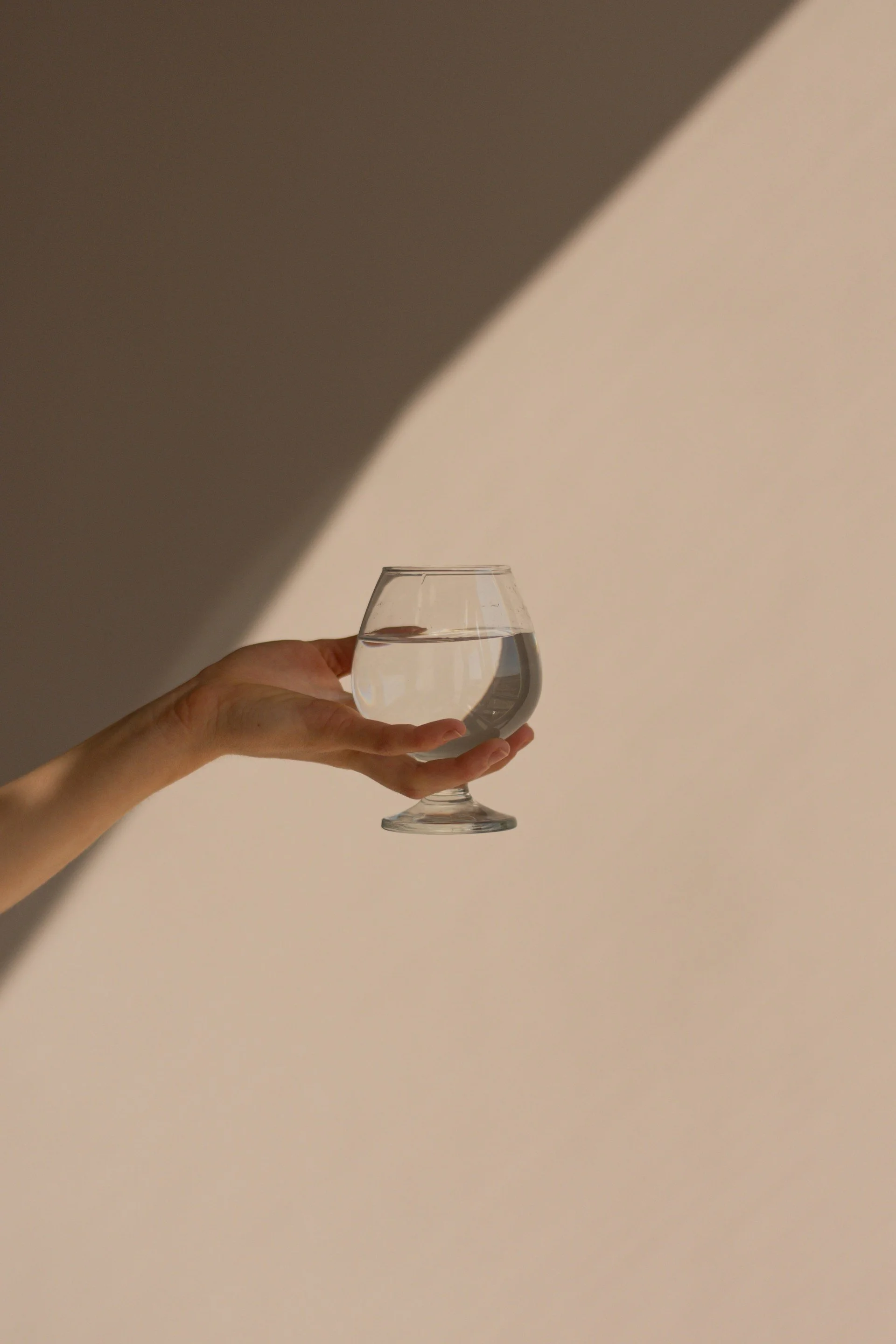 Person holding a glass of water against a beige background with dramatic shadow and lighting.