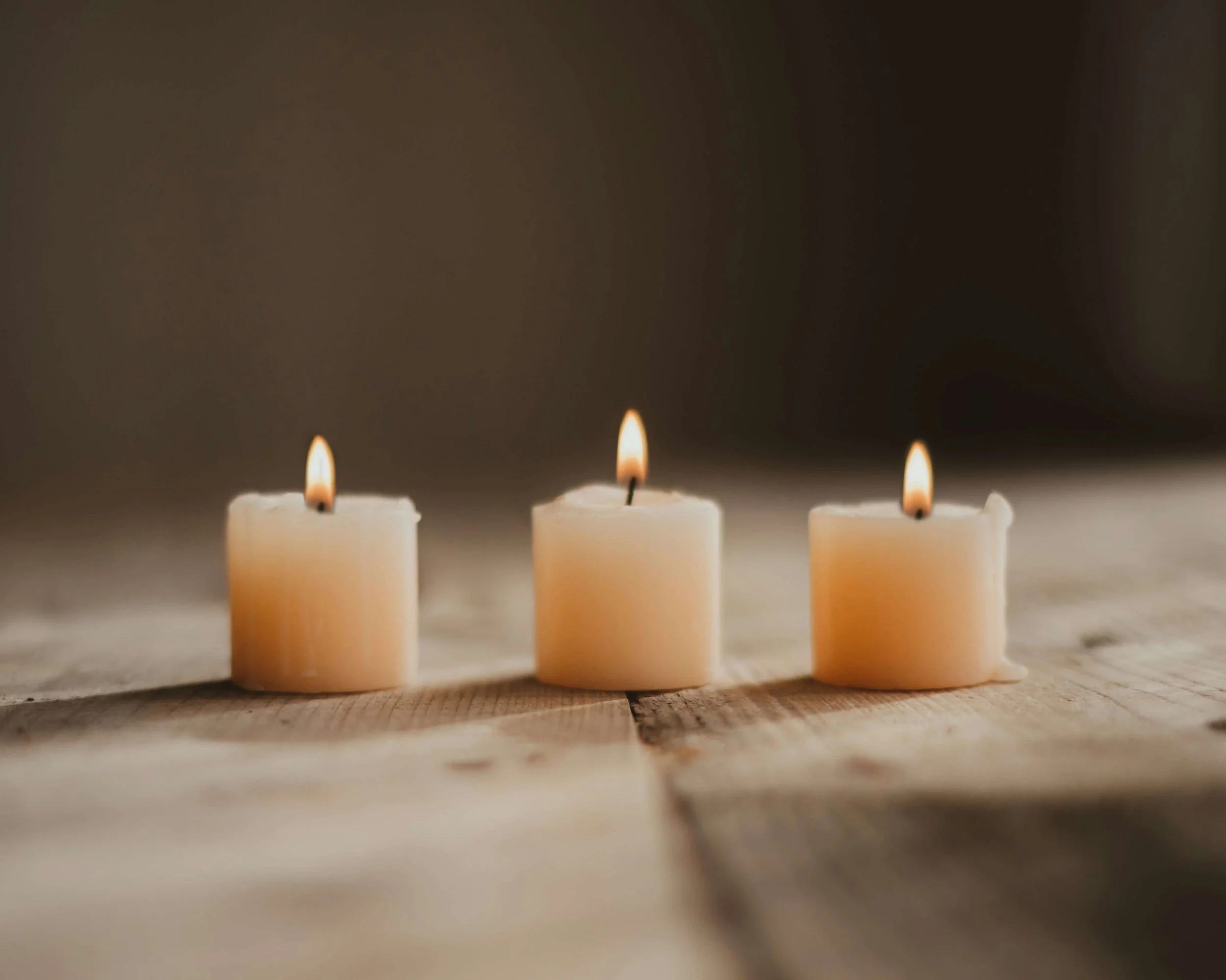 Three lit white birthday candles on a wooden surface with a blurred dark background.