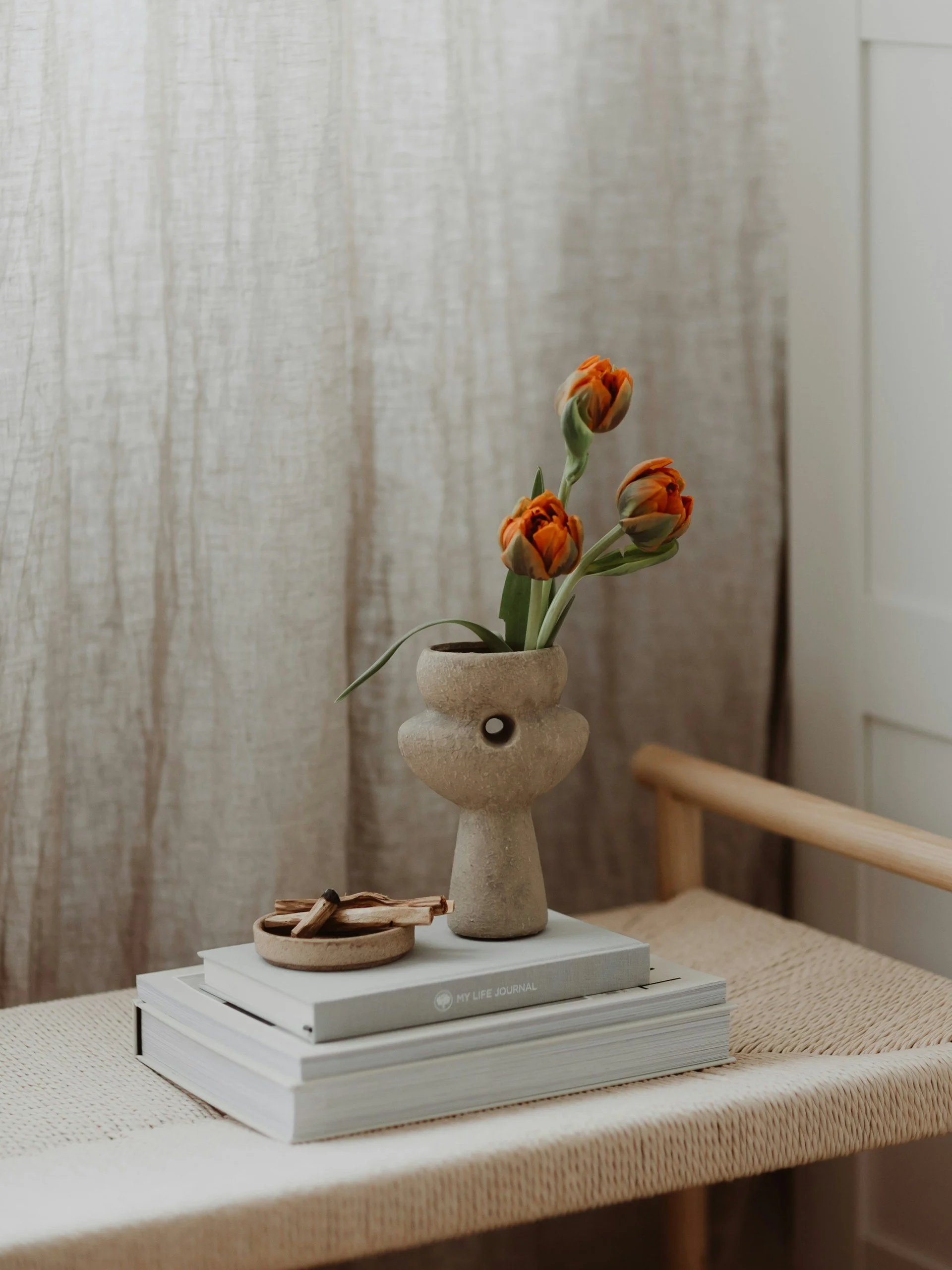 A beige ceramic vase with a face shape holds orange tulips on top of two stacked books, one titled 'My Life Journal', placed on a woven beige table. A small round dish with wooden sticks rests beside the vase, with beige curtains and white wall in the background.