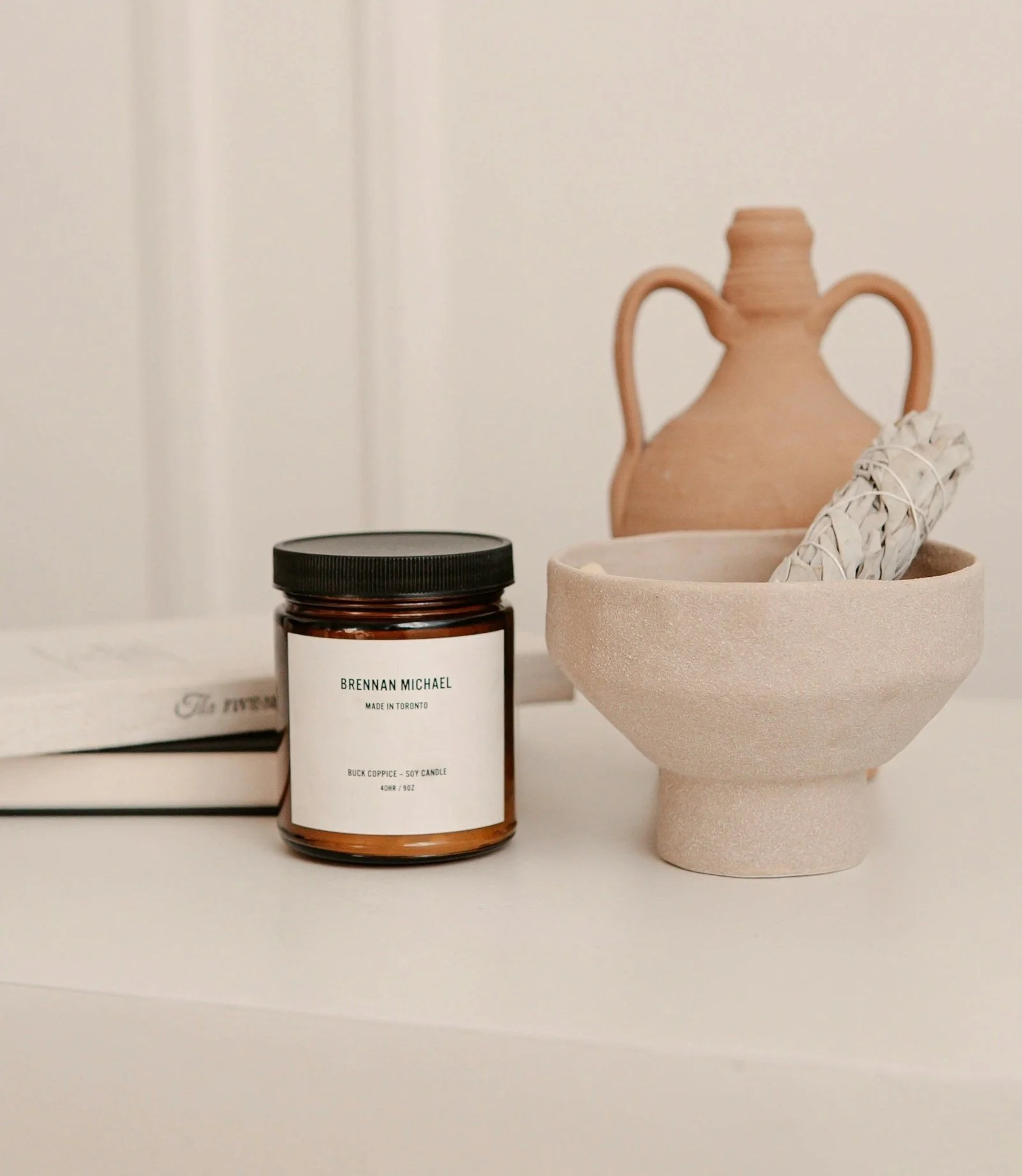 Decorative pottery vase, a beige ceramic bowl with a bundle of dried sage, and a brown candle in a jar with a black lid on a white surface against a neutral background.