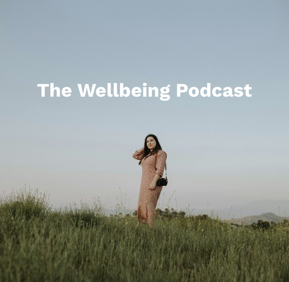 A woman standing in a grassy field with mountains in the background, holding a small purse, with the text 'The Wellbeing Podcast' overlaid on the sky.