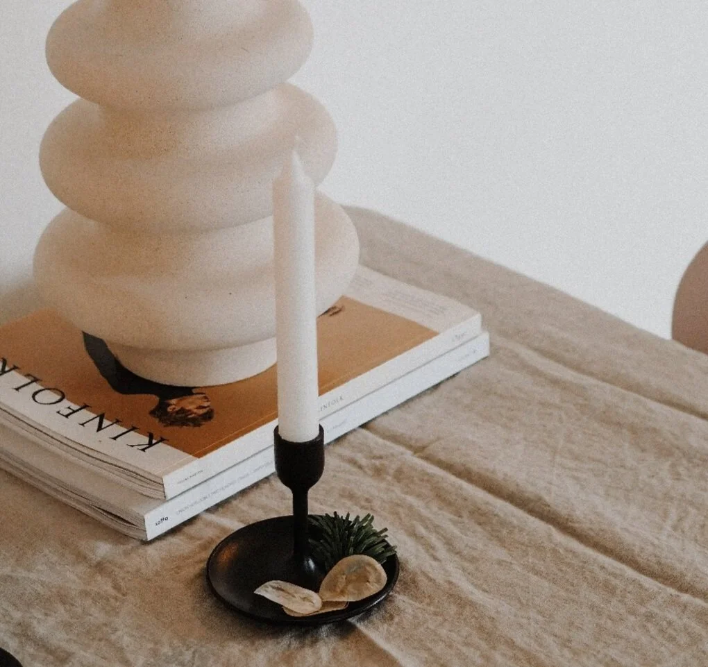 A black candle holder with a white candle, decorated with small rocks and a green plant, on a wooden surface with a magazine and abstract sculpture in the background.
