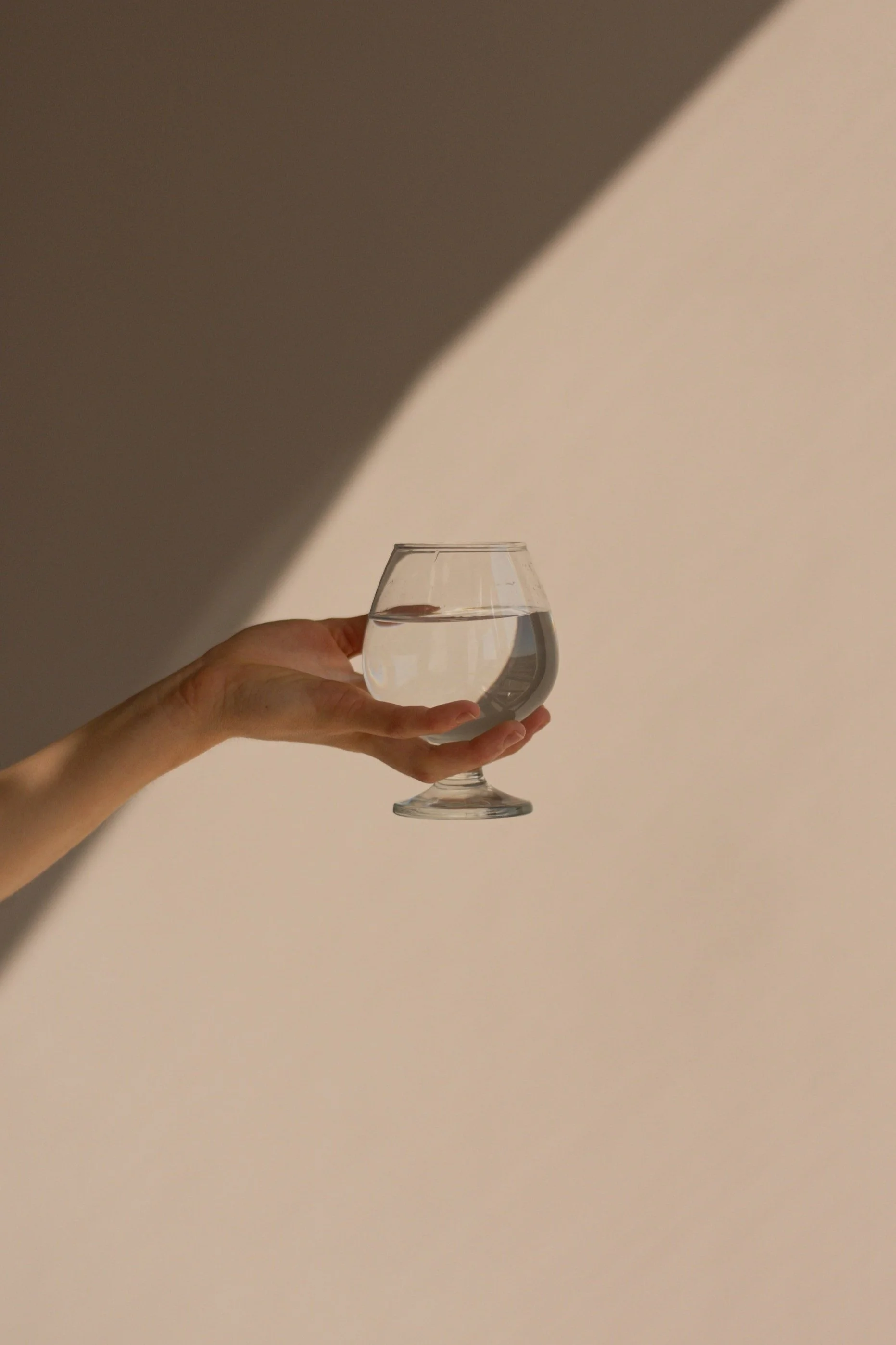 A person's hand holding a glass of water against a background with light and shadow.