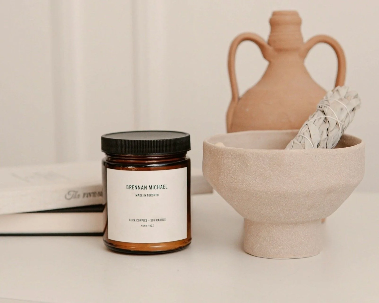 A ceramic bowl with a bundle of sage, a candle jar labeled Brendan Michael, and a brown ceramic vase on a white surface