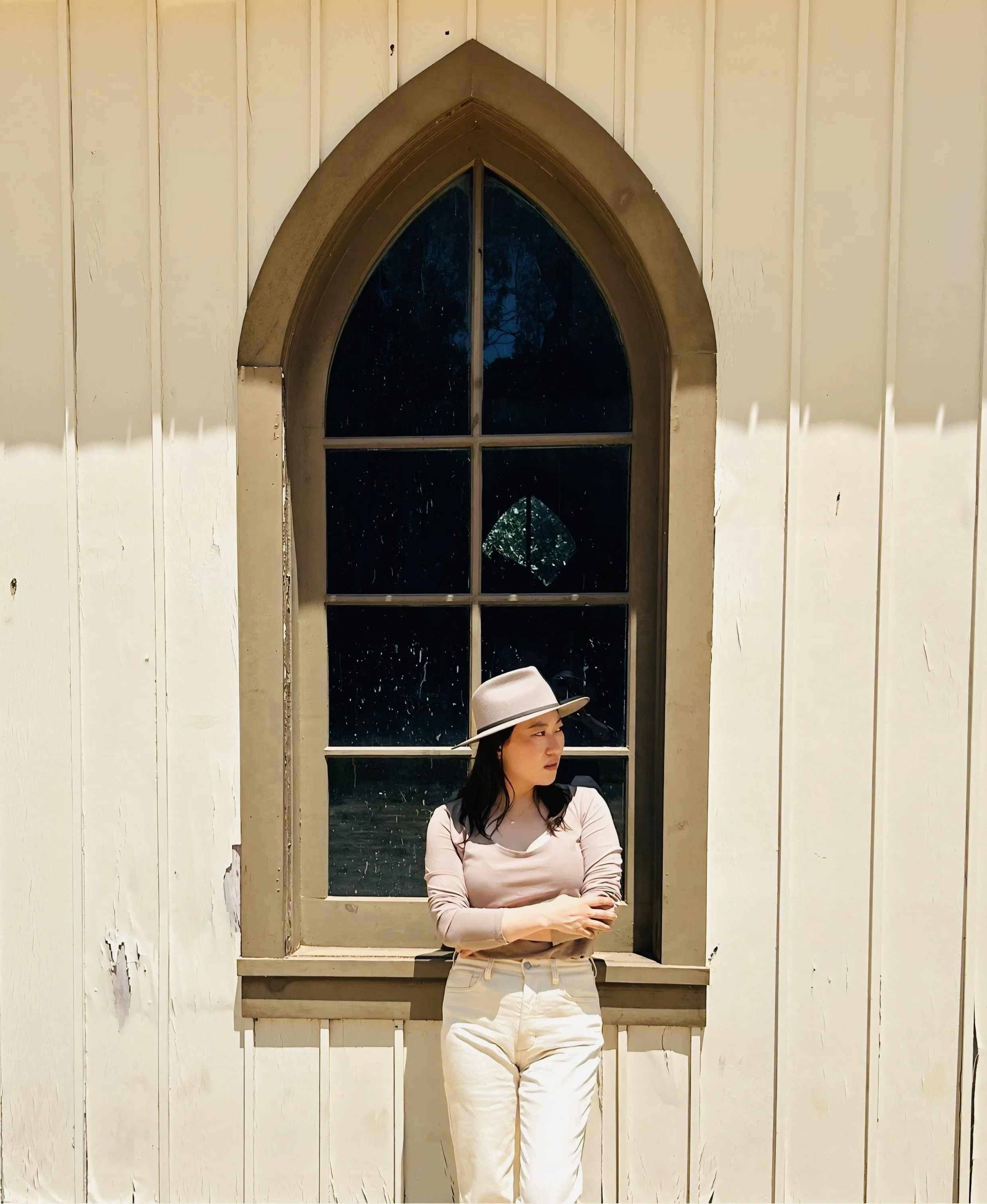 A woman standing in front of a gothic arched window on a white wooden wall, wearing a beige hat, a light long-sleeved top, and cream-colored pants.