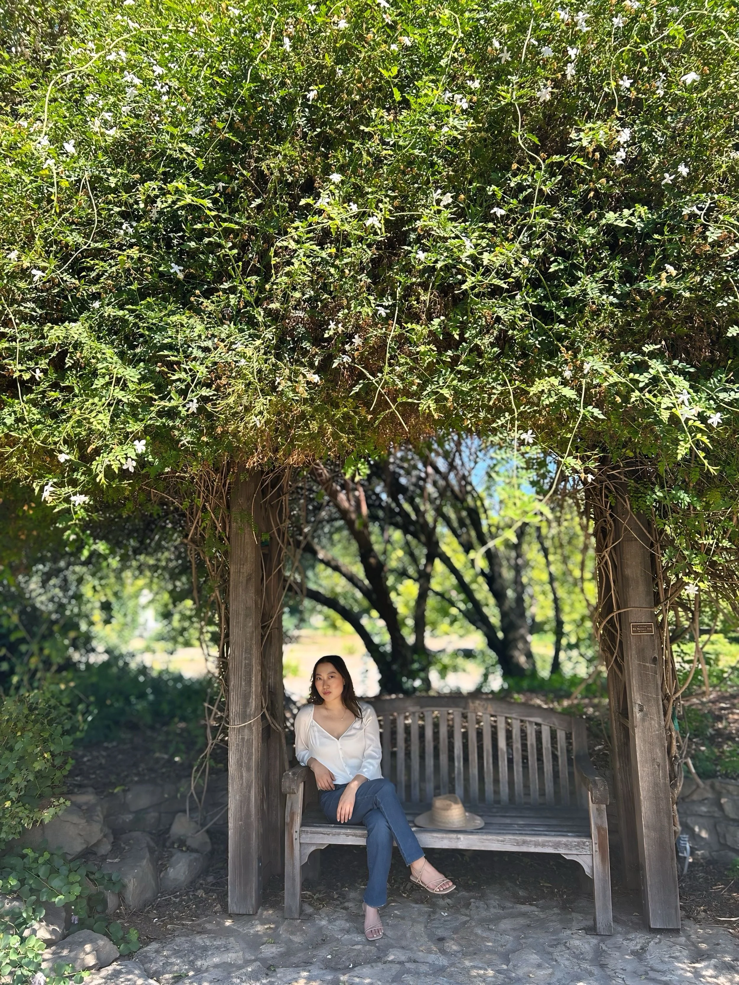 A woman sitting on a wooden bench under a leafy, vine-covered arbor in a lush garden.