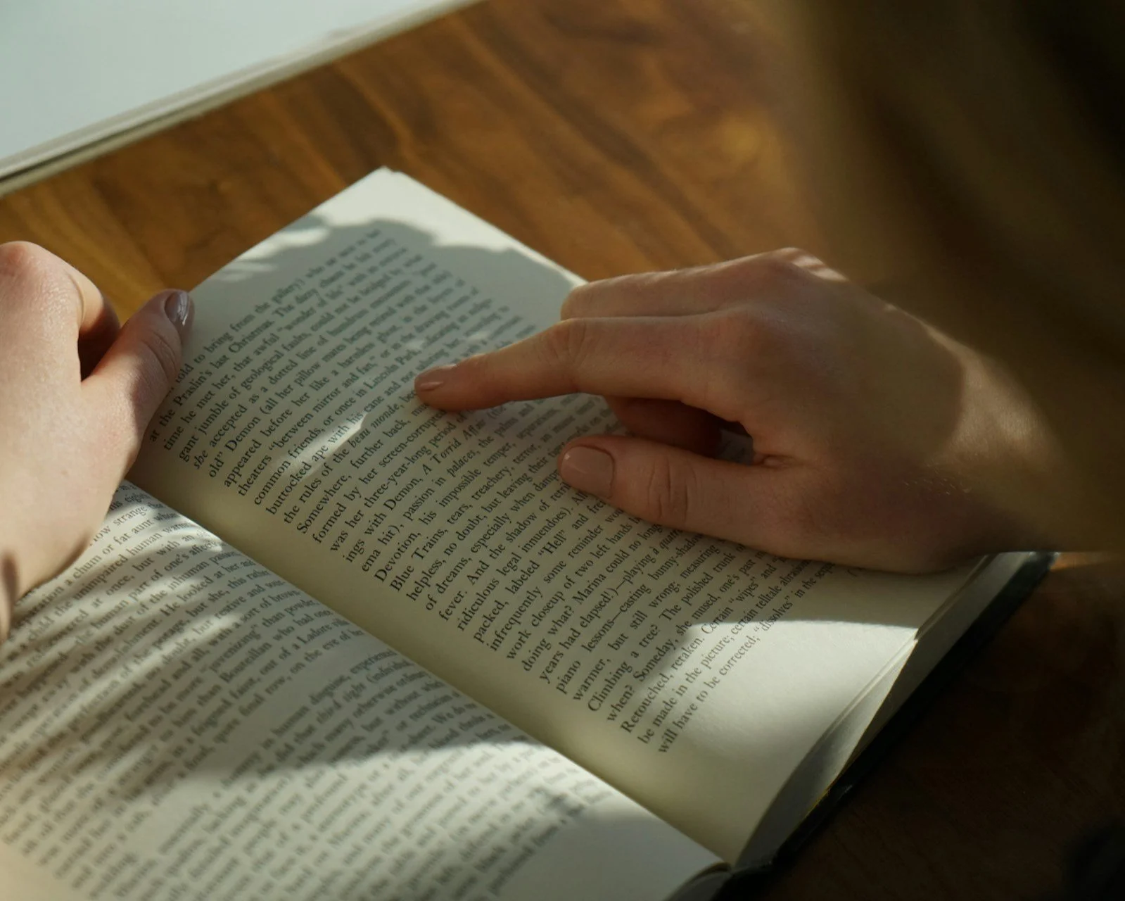 A person with light skin holding an open book, turning a page with their left hand, while sunlight illuminates the page on a wooden surface.
