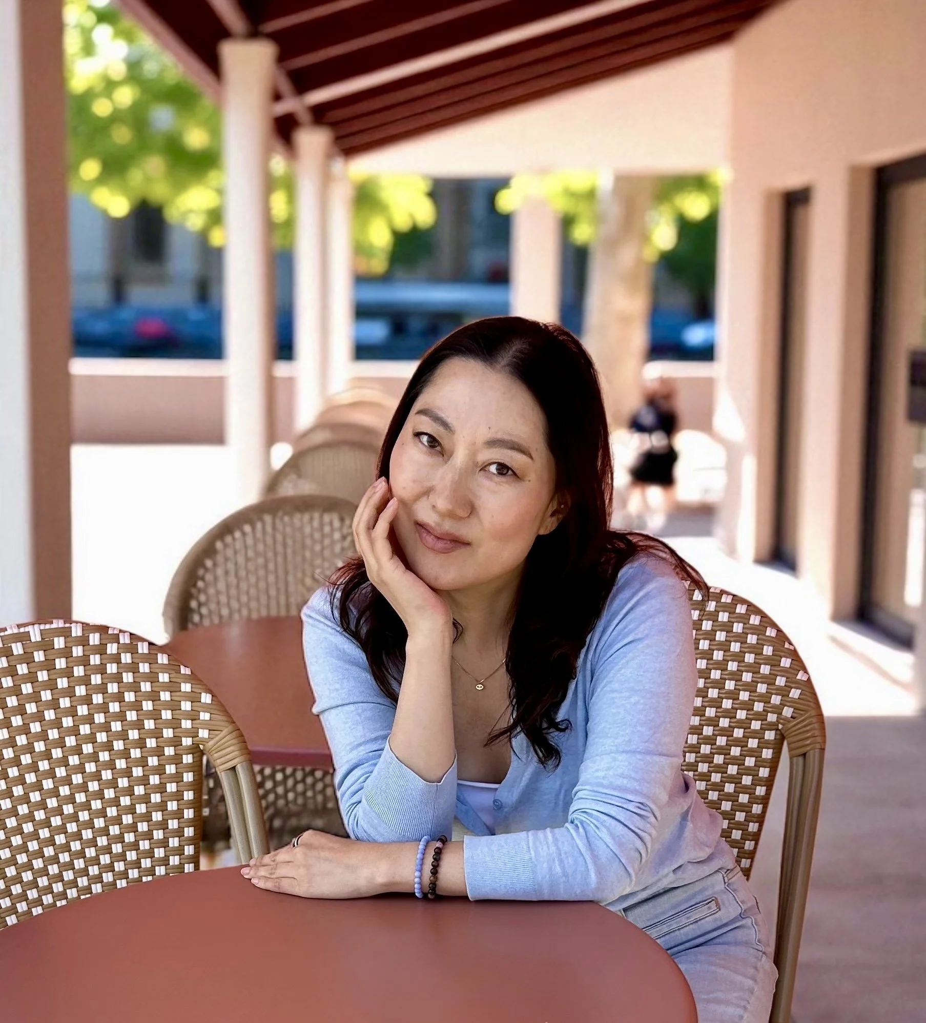 A woman with dark hair and light clothing sitting at an outdoor cafe table, resting her head on her hand, with a blurred background of trees and a person walking.