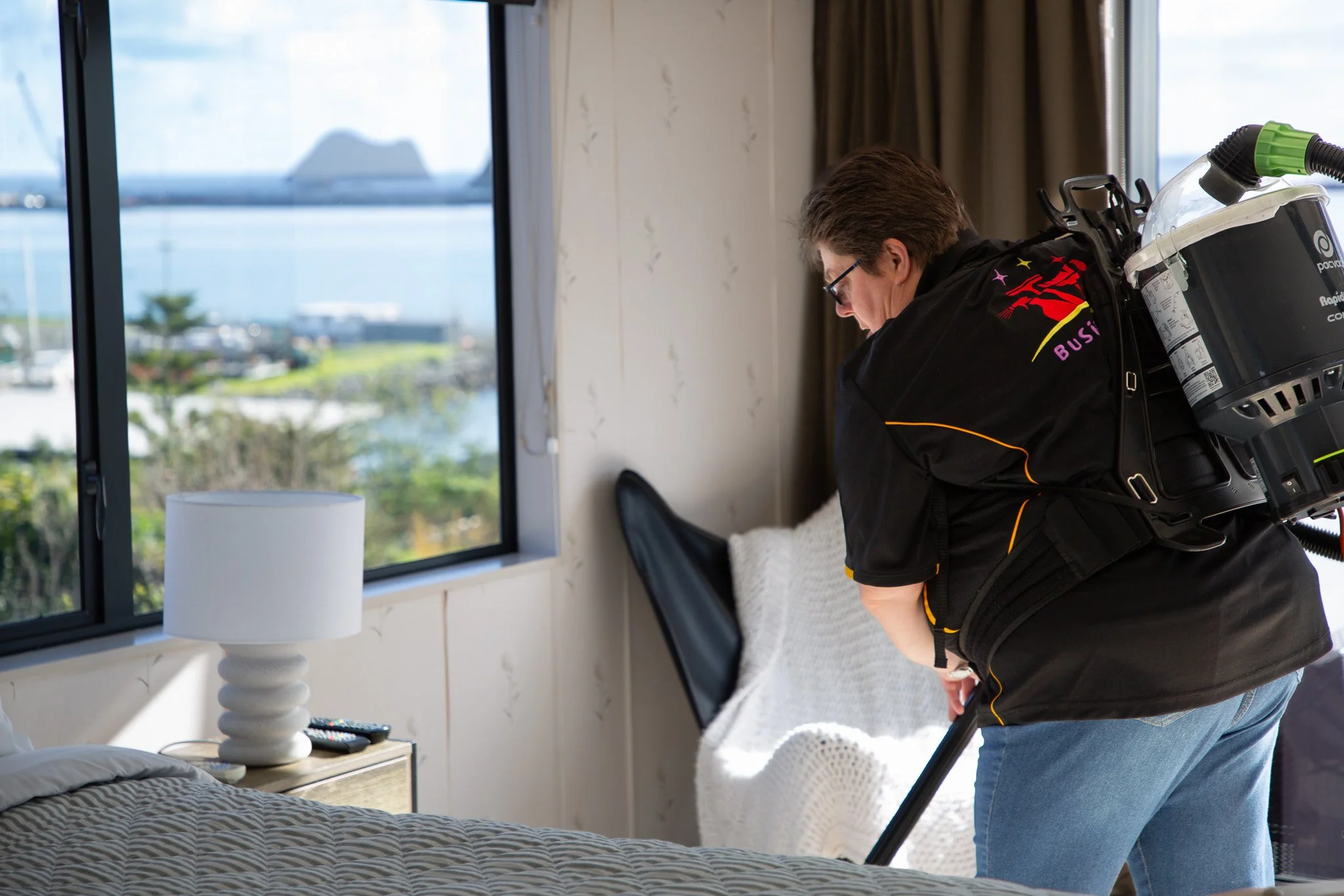 Person cleaning or vacuuming a bed in a hotel room with a window showing an outdoor view.