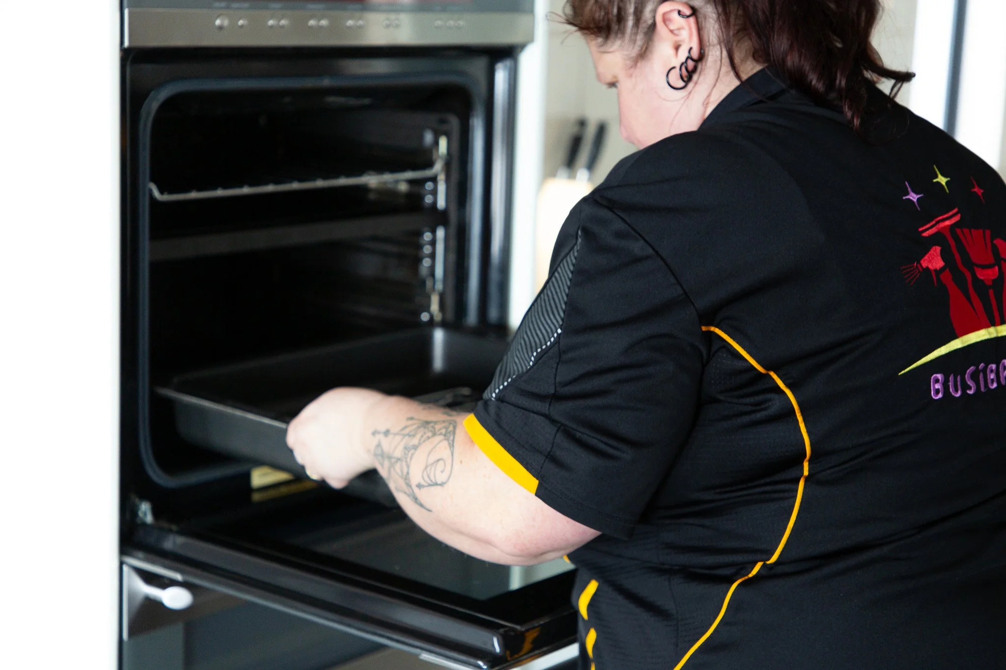 A woman with tattoos on her right arm placing a tray into an empty oven.