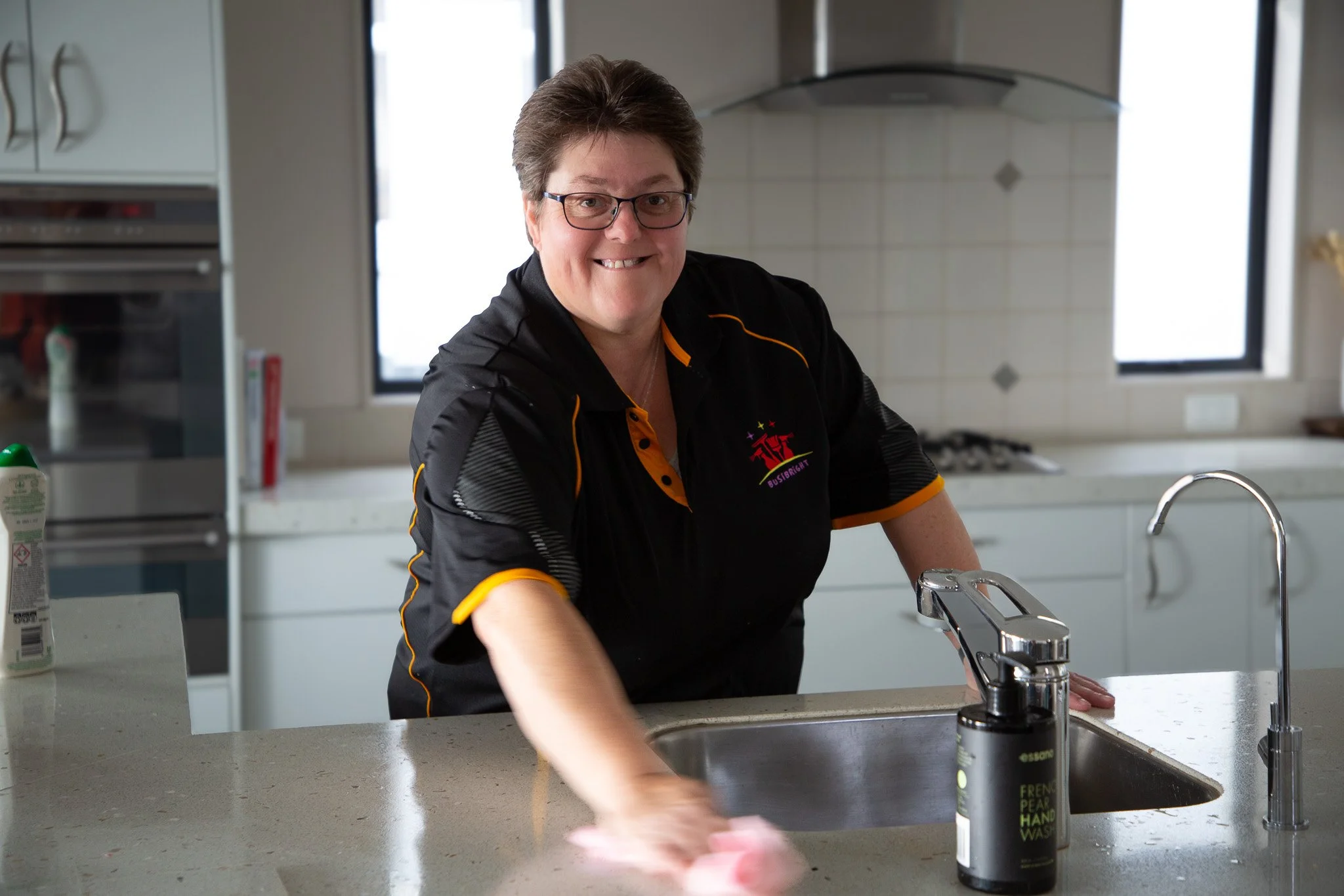 Woman cleaning a kitchen countertop with a pink cloth, wearing glasses and a black sports jersey with yellow accents.