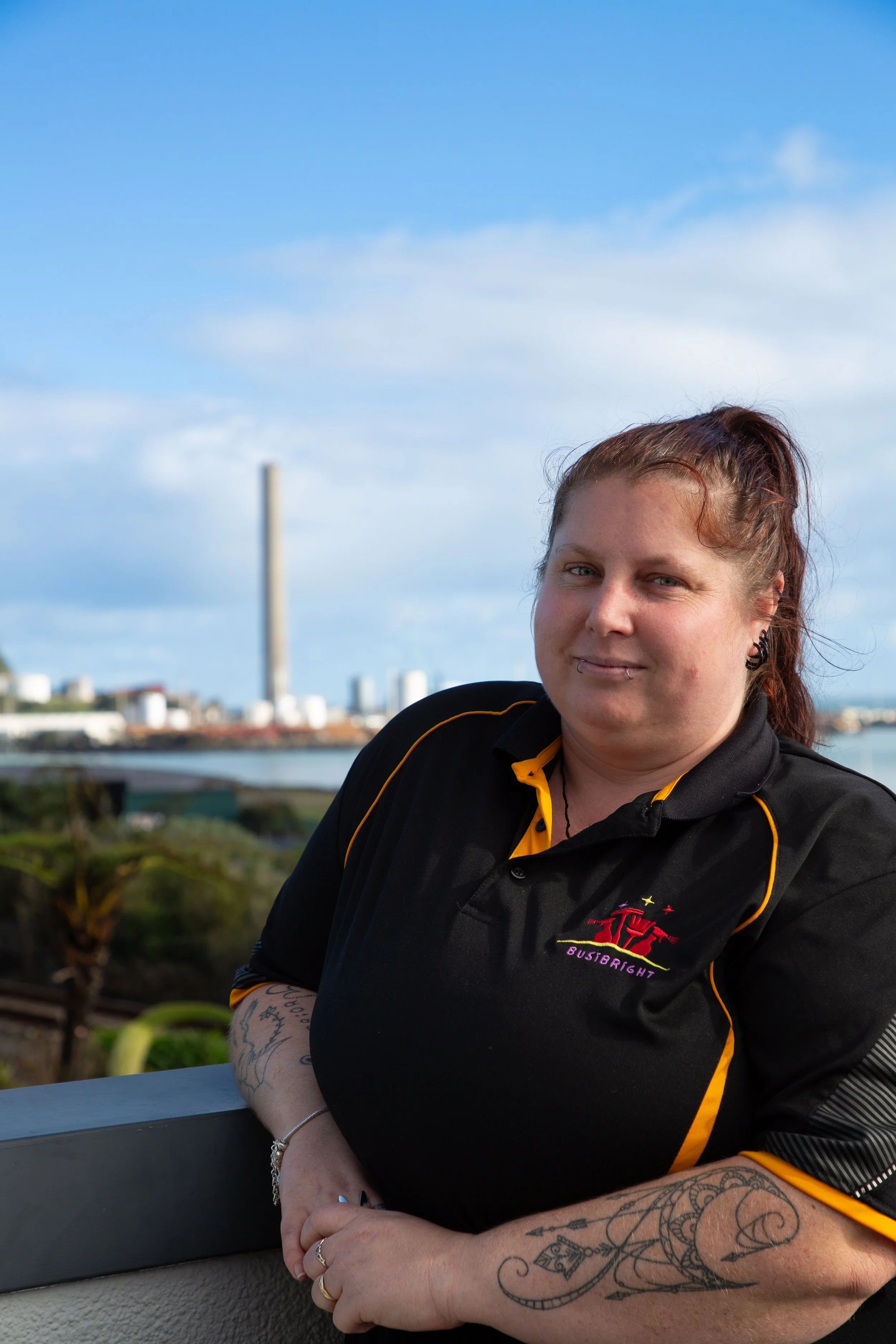 A woman leaning on a railing outdoors, with a city skyline and body of water in the background, wearing a black and yellow sports shirt.