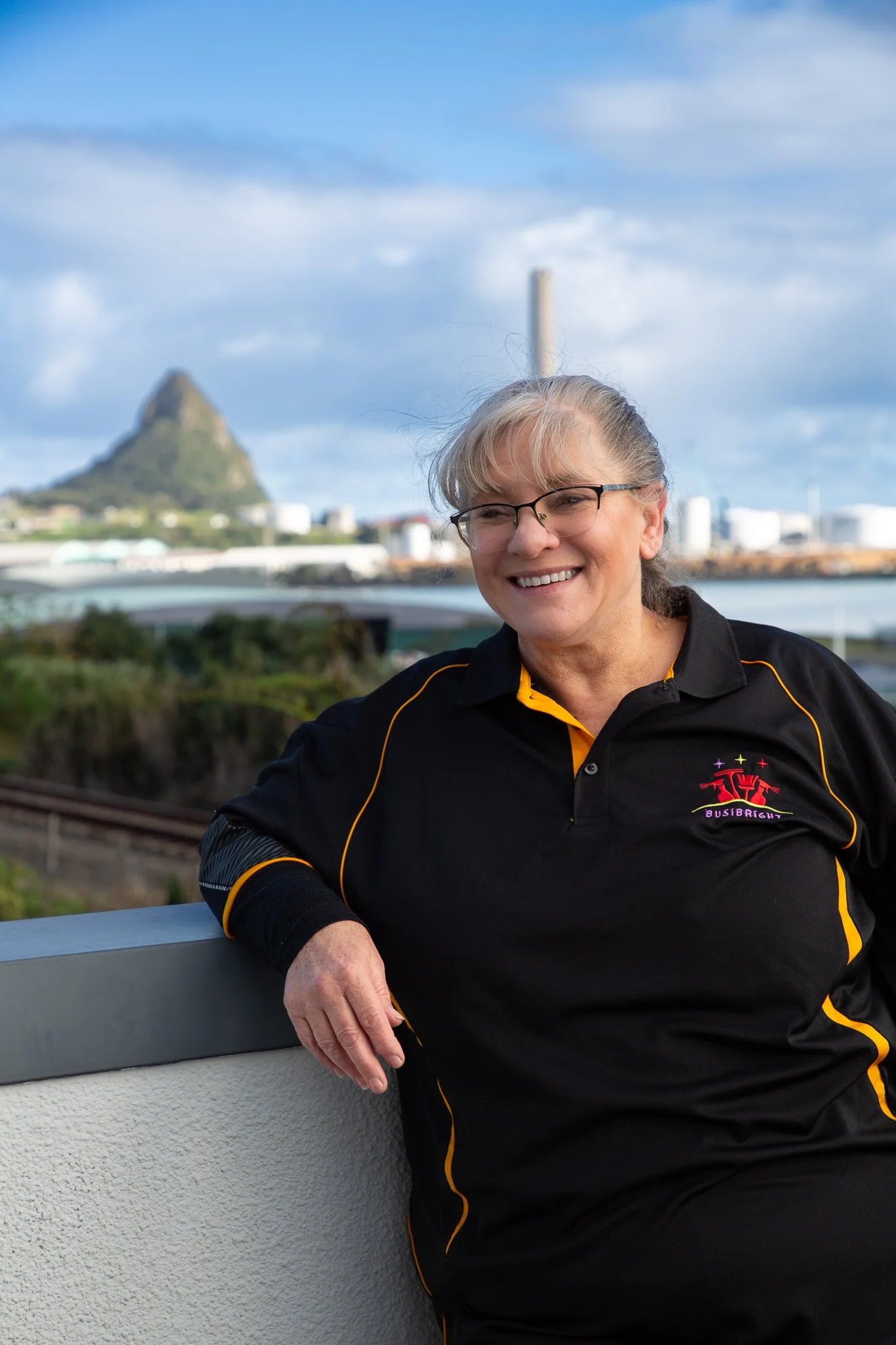 A smiling woman with gray hair and glasses wearing a black sports shirt with yellow accents, standing outdoors near a railing at a distance, with a mountain and industrial buildings in the background.