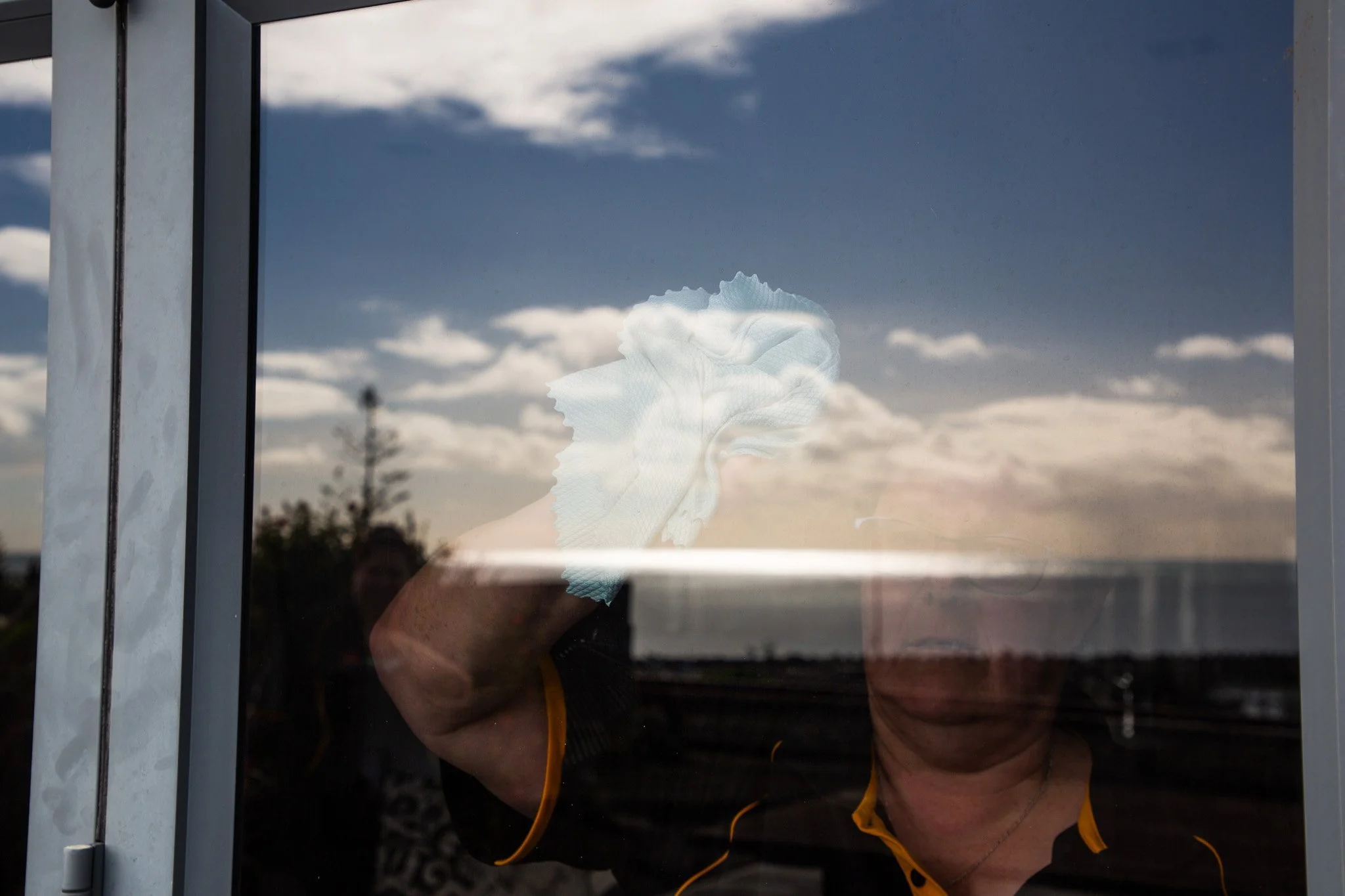 Person cleaning window glass with a tissue or cloth, reflection of person and outdoor sky with clouds visible on the glass