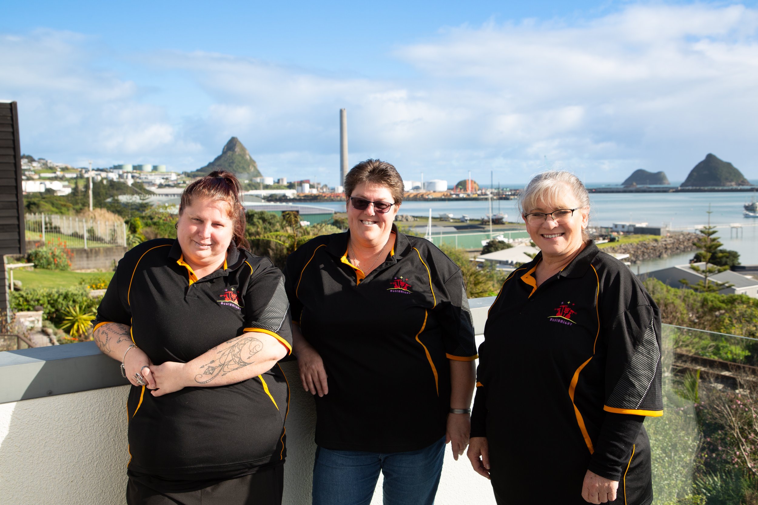 Three women wearing black sports shirts with yellow accents, standing outdoors with a coastal cityscape and mountains in the background, smiling at the camera.