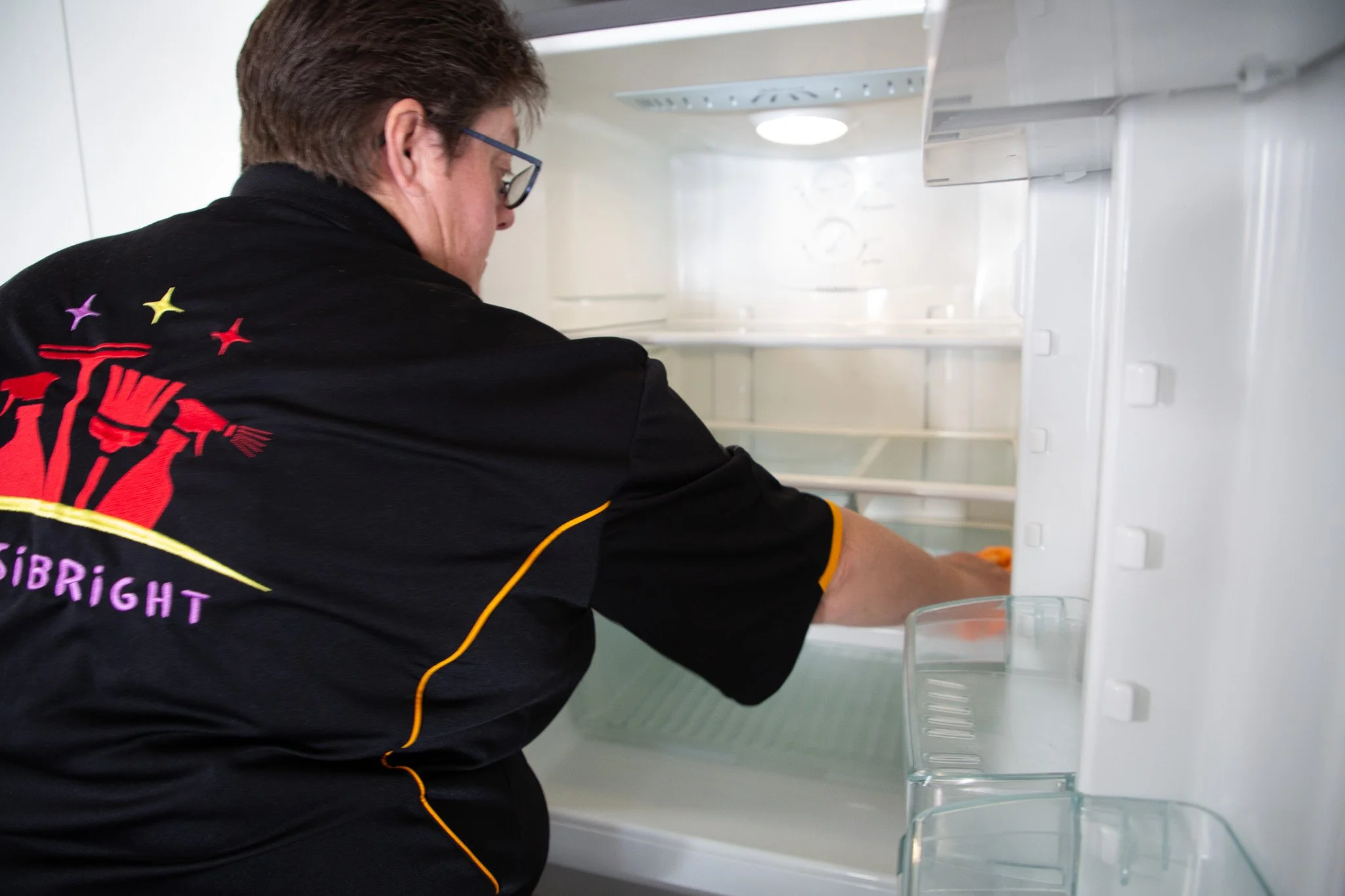 Person cleaning an empty refrigerator shelf inside a white fridge.