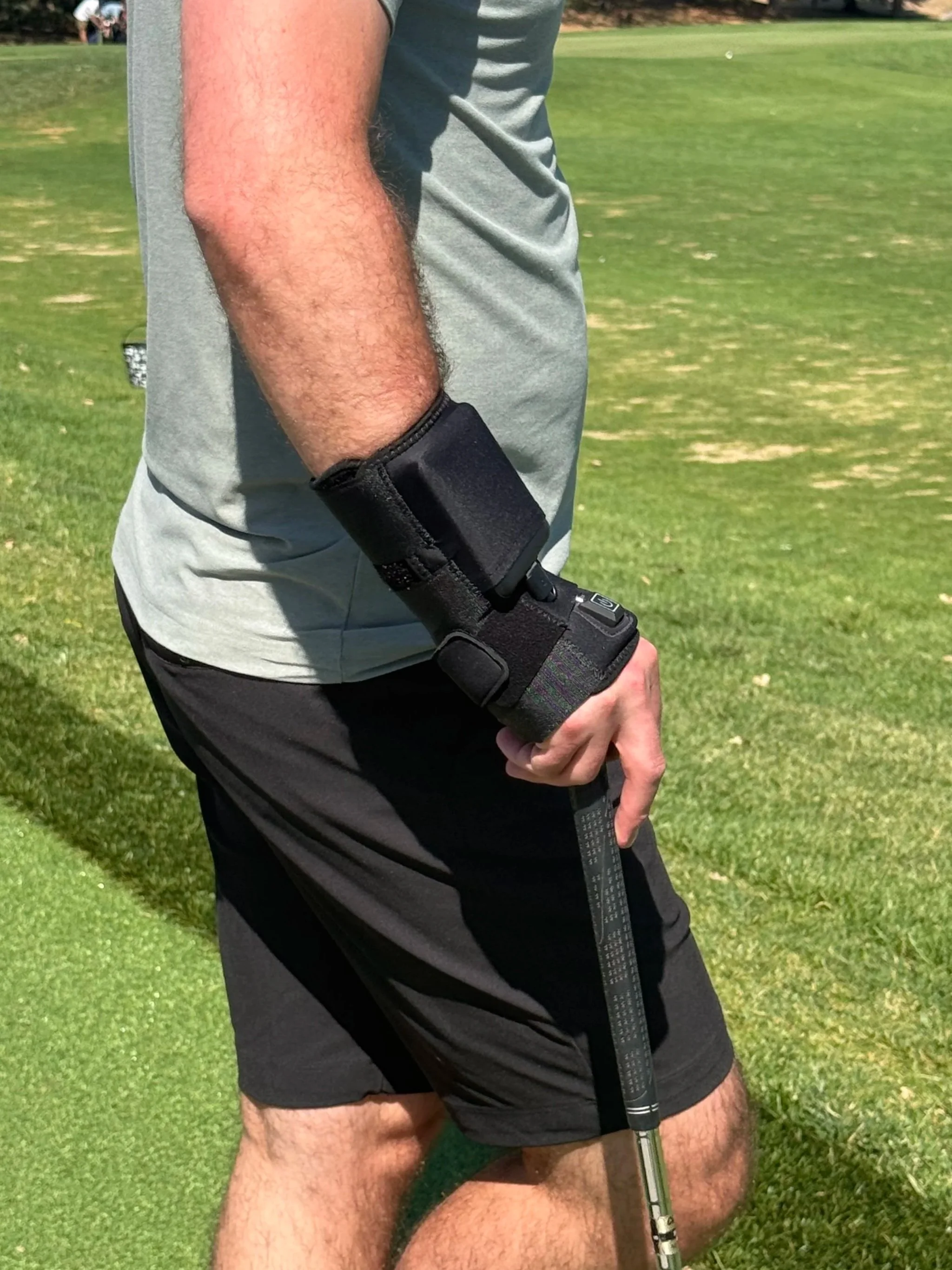 A person dressed in athletic clothing standing on a grassy field, holding a golf club with a thermera heated wrist brace on their right arm.